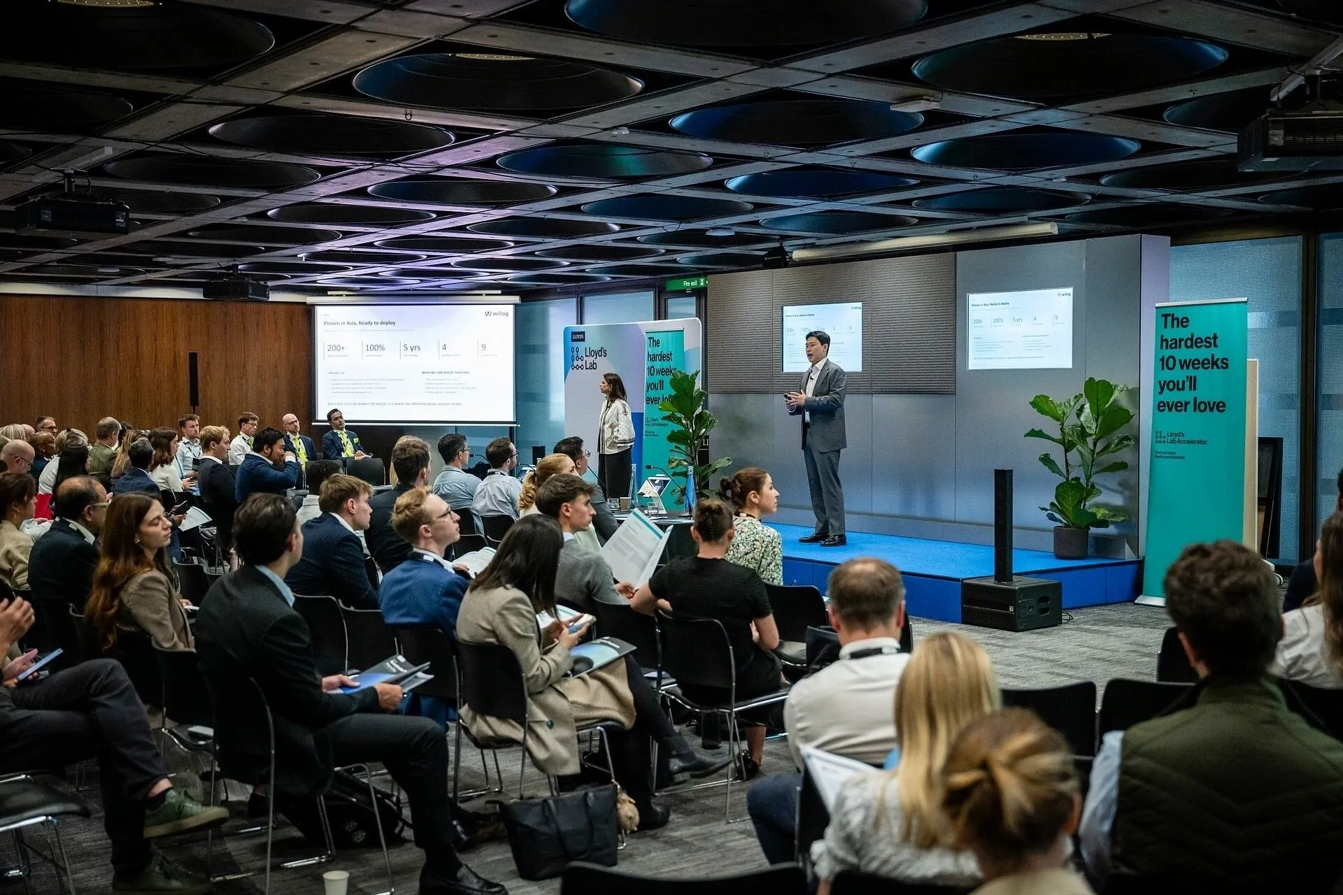 Audience seated at a business presentation with two speakers on a blue stage and Lloyds Bank banners.
