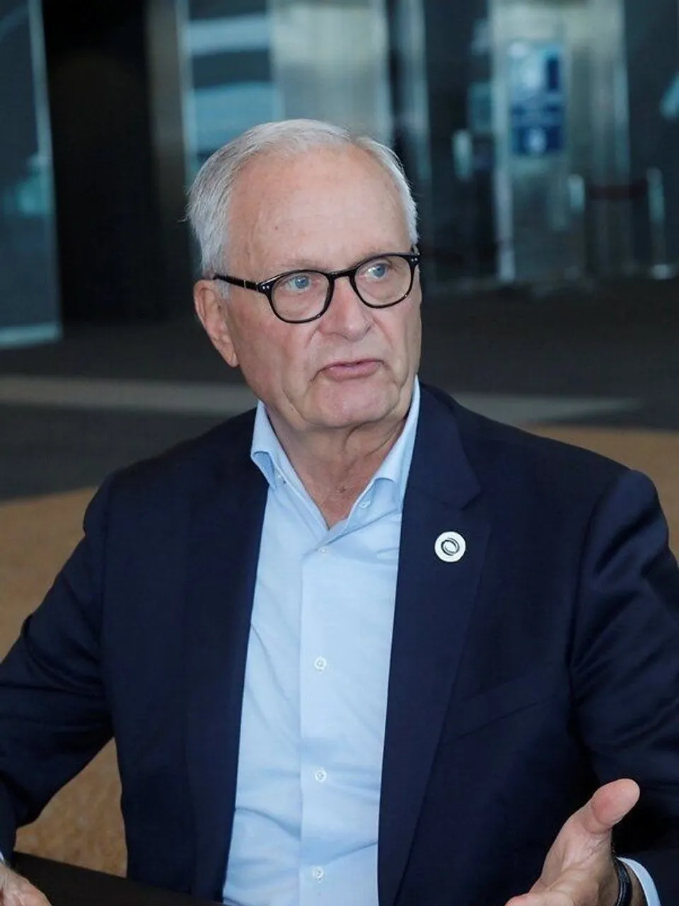 An older man in glasses and a navy suit gestures while seated indoors.
