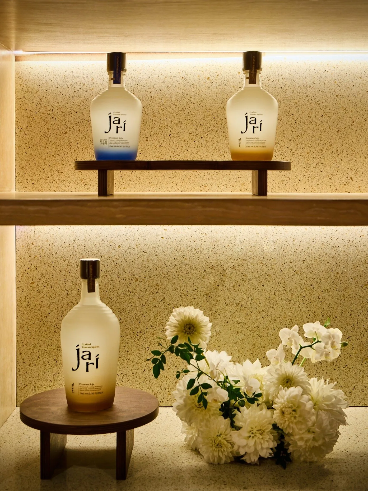 Three bottles of Jari premium soju on illuminated wooden shelves beside white flowers
