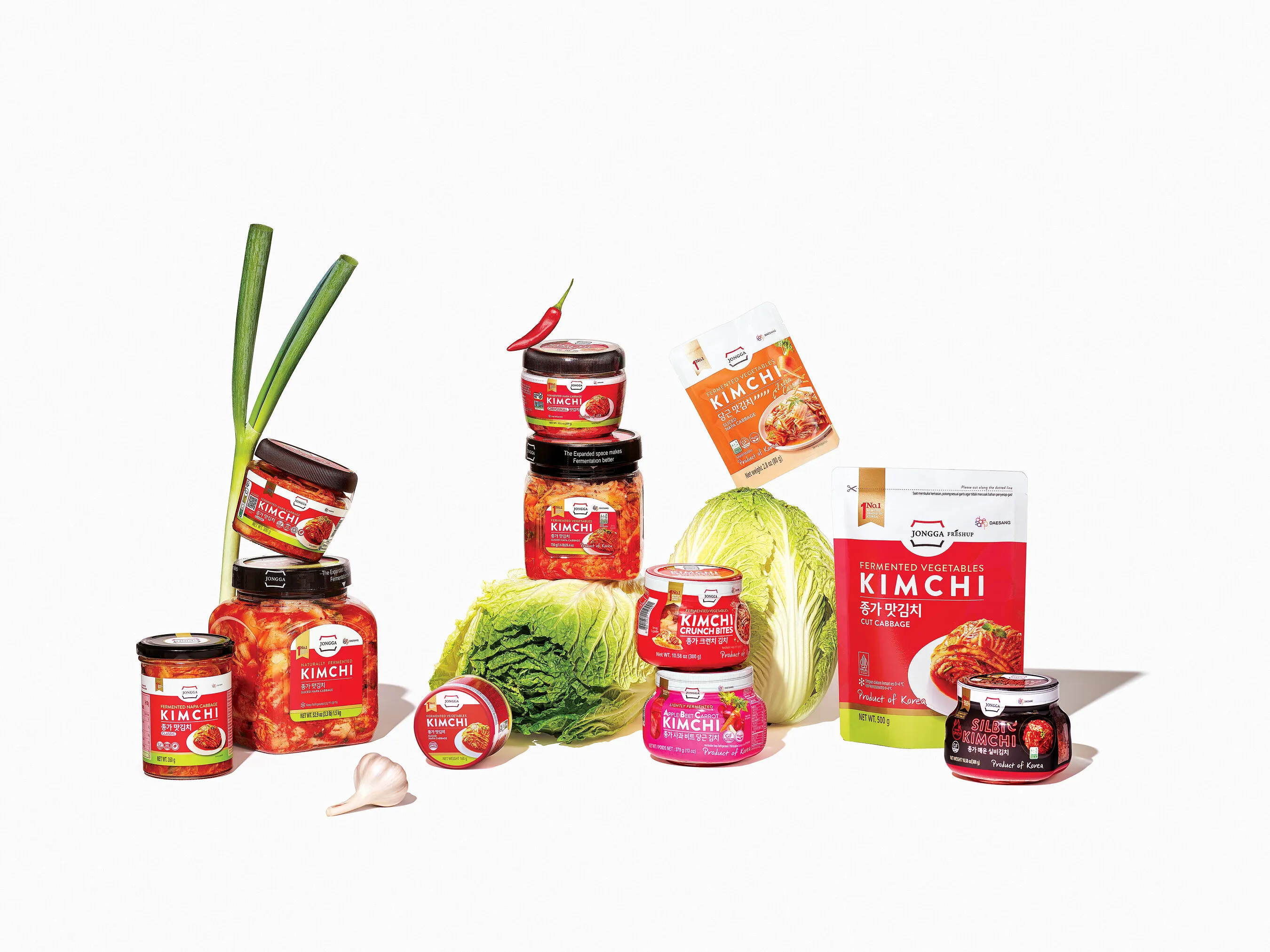 Assorted kimchi jars, cans and pouches arranged with napa cabbage, scallions, garlic and a red chili pepper.