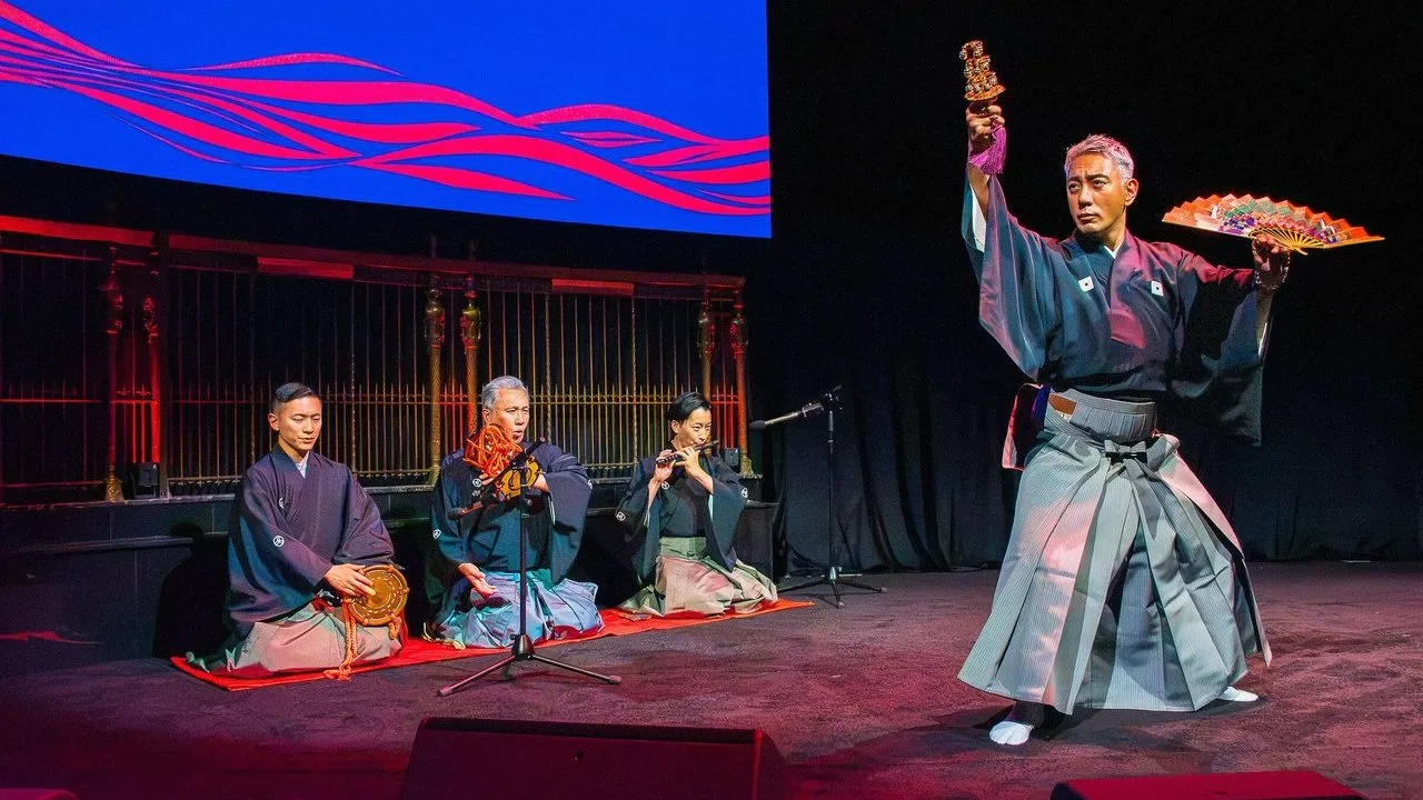 A performer in traditional Japanese dress holds a fan and ornament beside seated musicians on a stage.