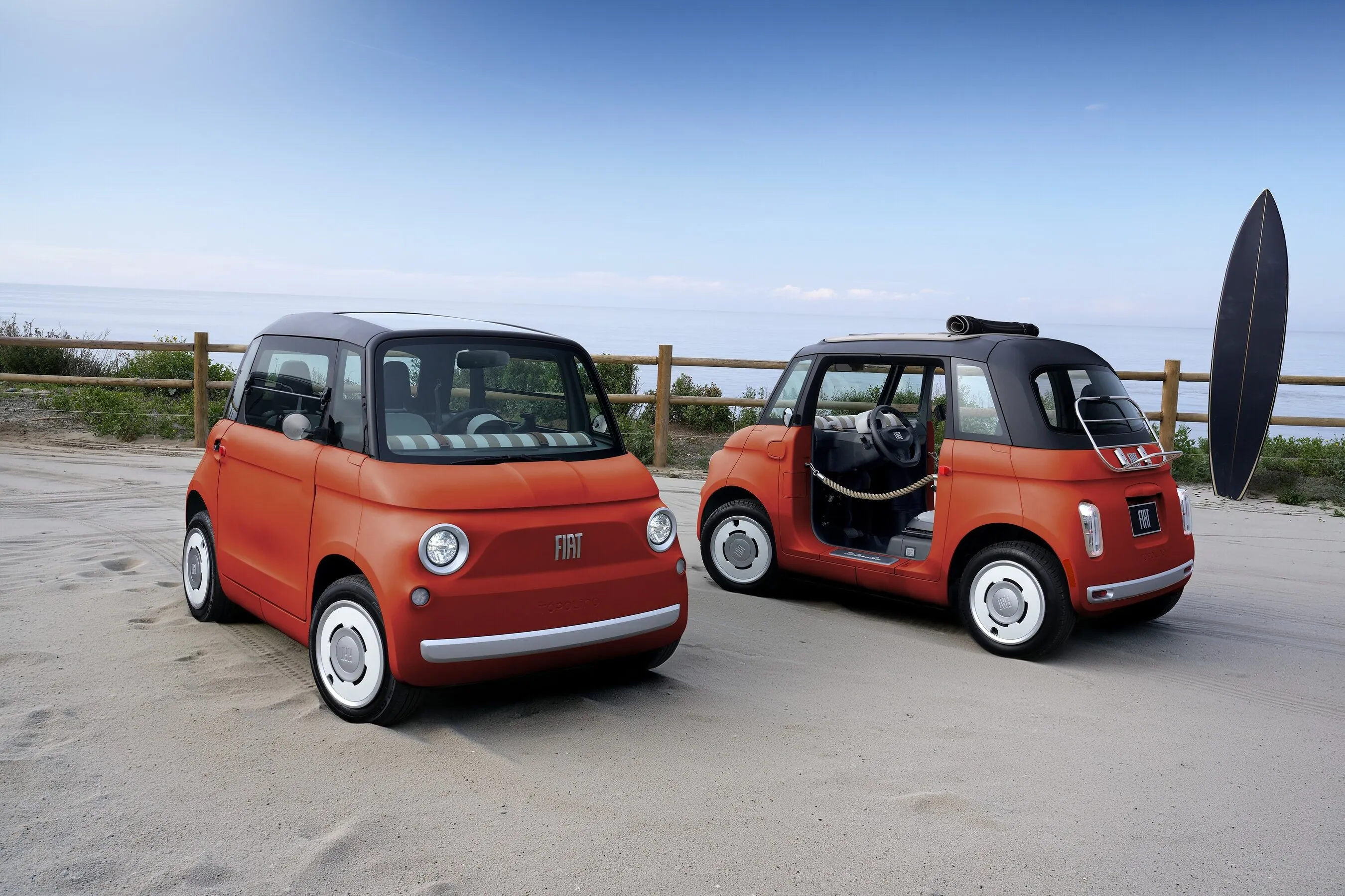 Two orange Fiat Topolino cars parked by the beach, one with a surfboard mounted at the rear.