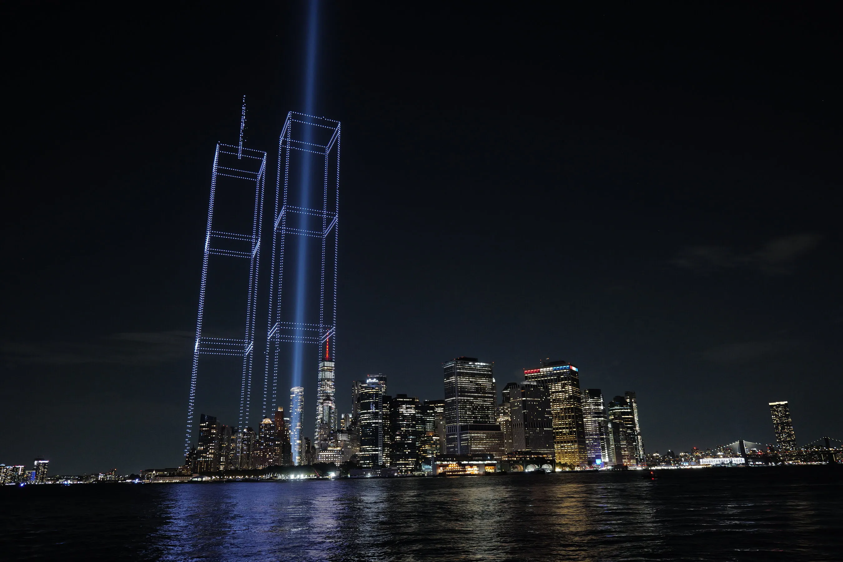 Blue beams and outlined Twin Towers rise above the illuminated Manhattan skyline at night.