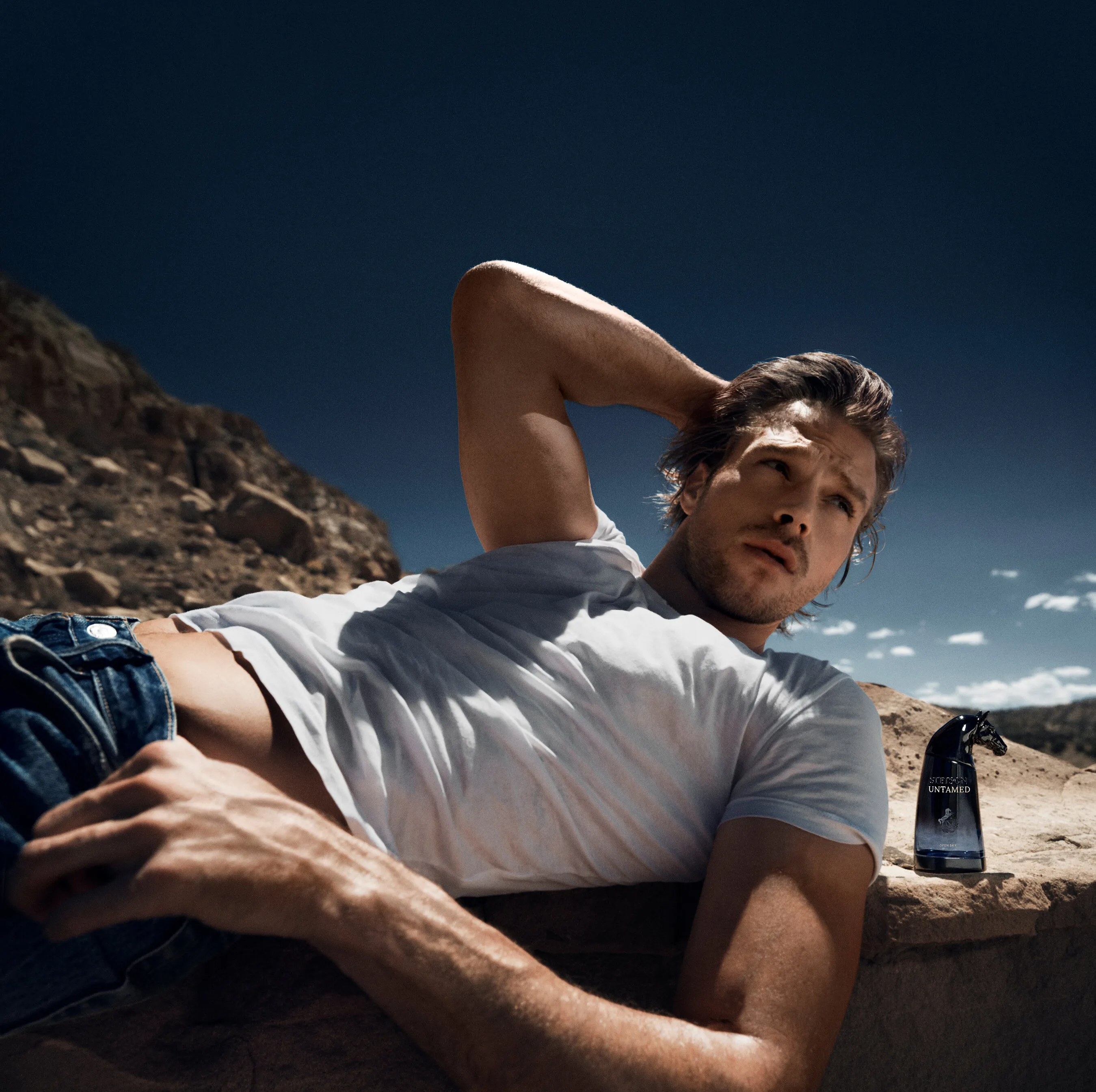 Matthew Noszka reclining in a white T-shirt and jeans beside a dark fragrance bottle in a rocky desert.