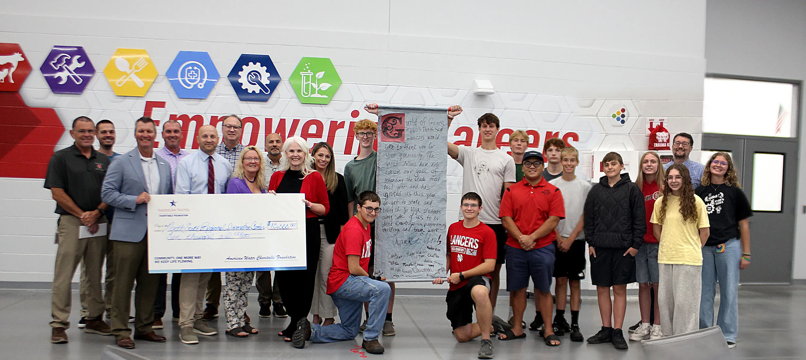 A group of students and adults hold a large check and signed banner in a school hallway.
