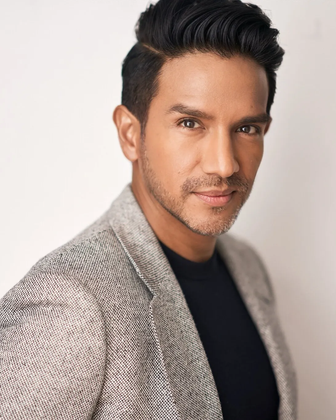 A man with styled dark hair and stubble wearing a gray blazer over a black shirt.