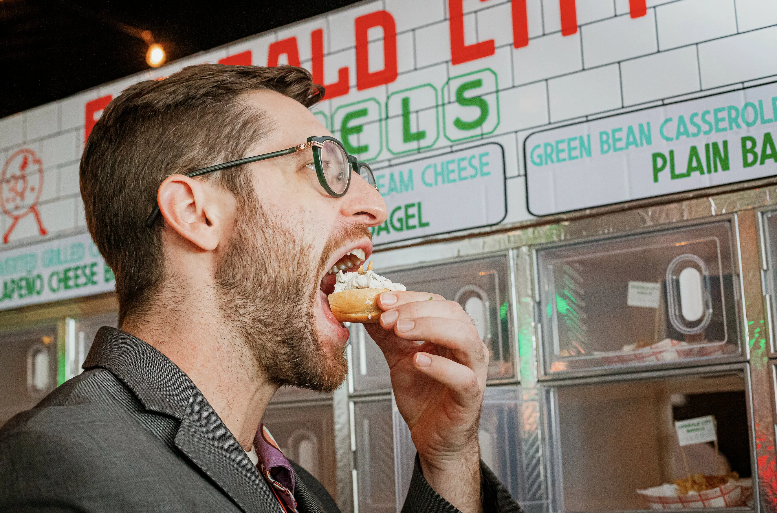 Sam Silverman eating a cream cheese bagel in front of a bagel display.