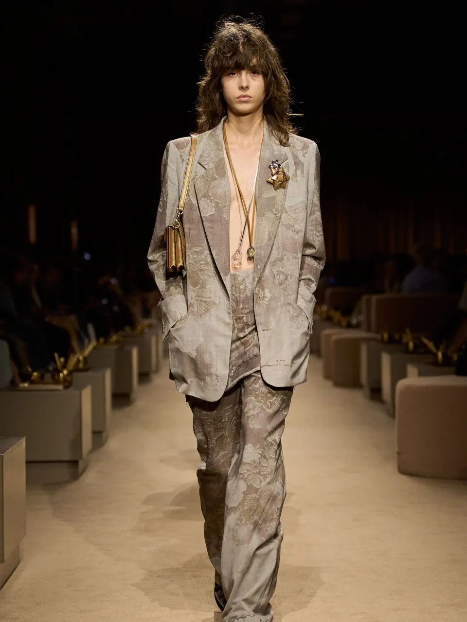 Model in a floral beige suit with layered necklaces and a shoulder bag walking a runway