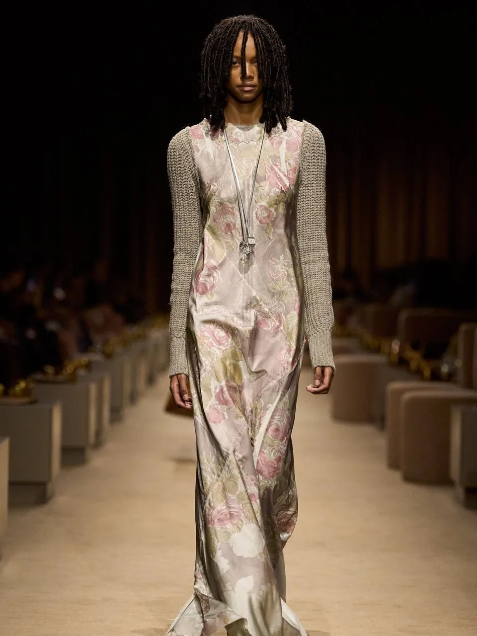 Model in a floral satin gown, knit sleeves and layered necklaces walking a runway.