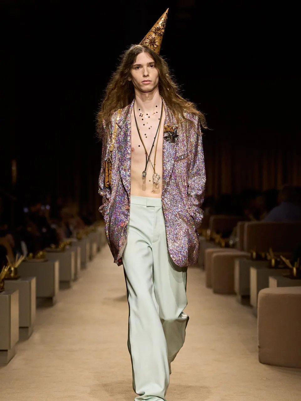 Model in a jeweled cone hat, metallic blazer and pale trousers walking a runway