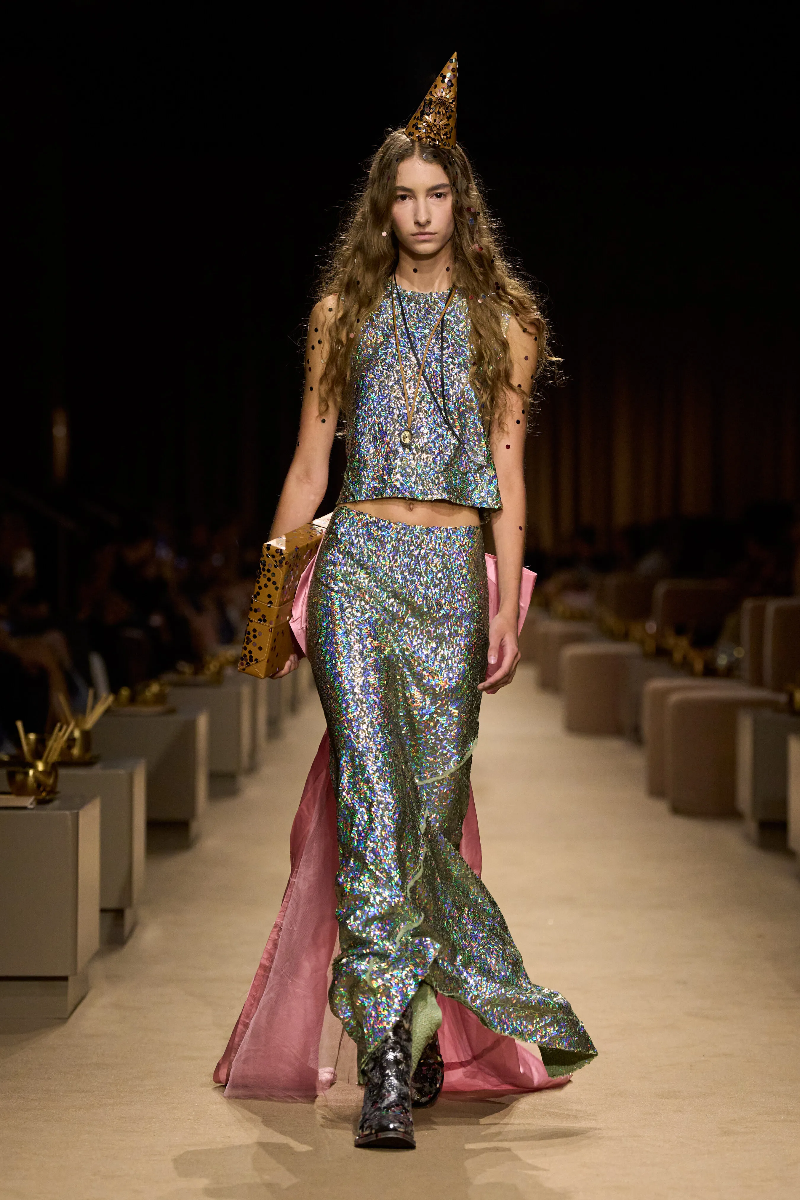 Model in a shimmering silver two-piece outfit, sheer polka-dot sleeves and a pointed hat on a runway.