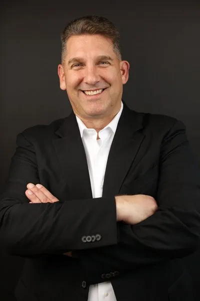 Erich Sanchack in a black suit and white shirt, smiling with arms crossed.