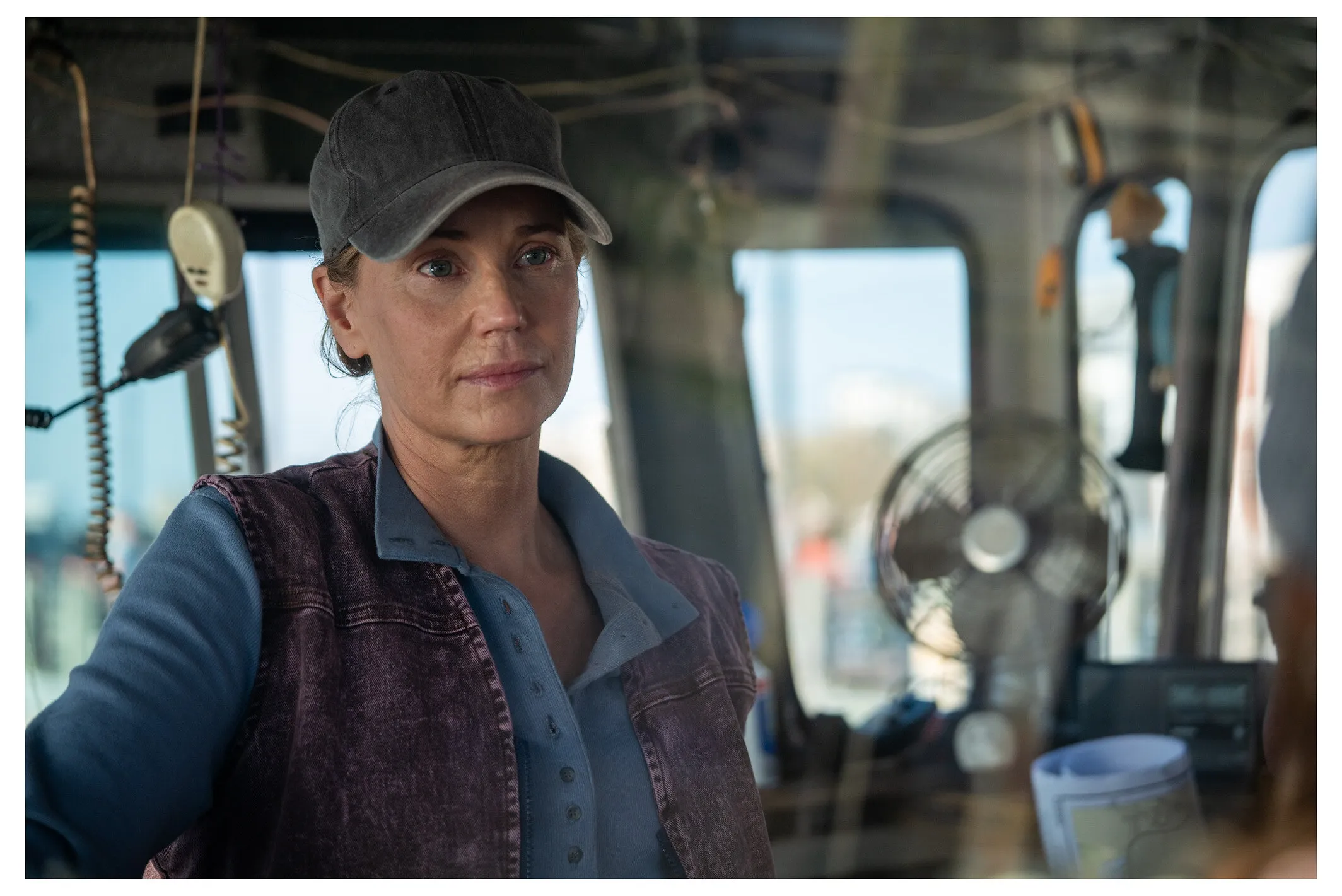 A woman in a cap sits at the controls inside a boat cabin.