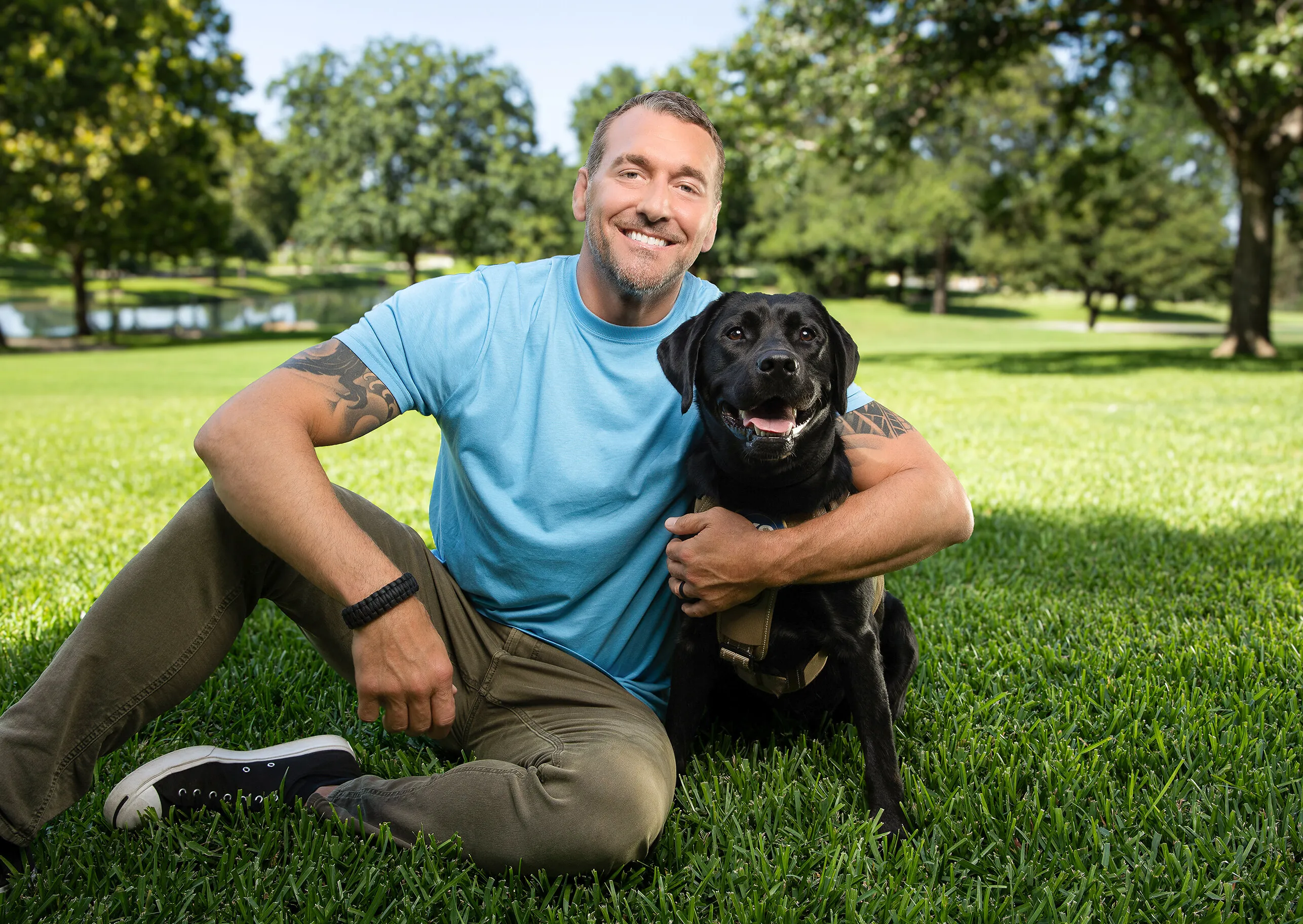 Brandon McMillan sits on grass holding a black Labrador retriever in a park.