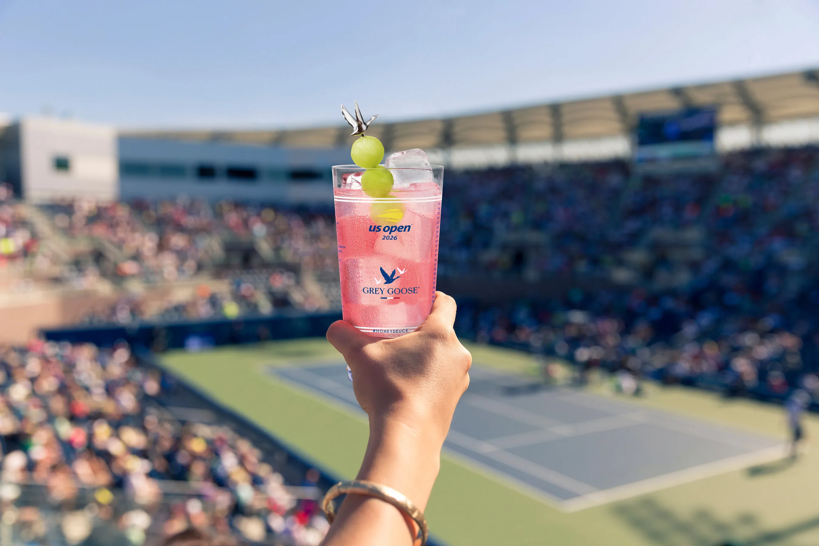 Hand holds a pink Honey Deuce cocktail garnished with melon balls above a crowded tennis stadium.