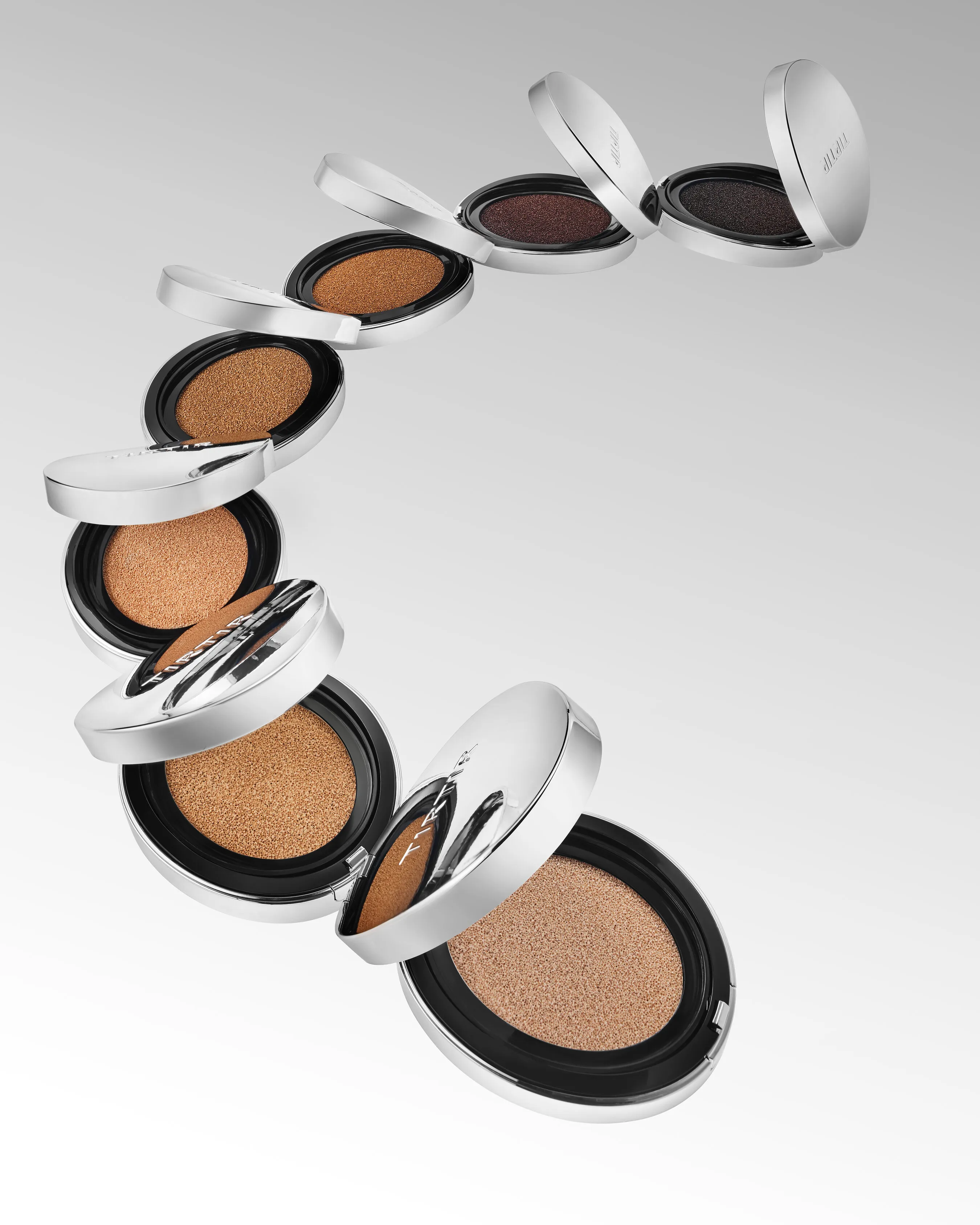 Open silver compacts containing cushion foundation in varied shades against a light gray background