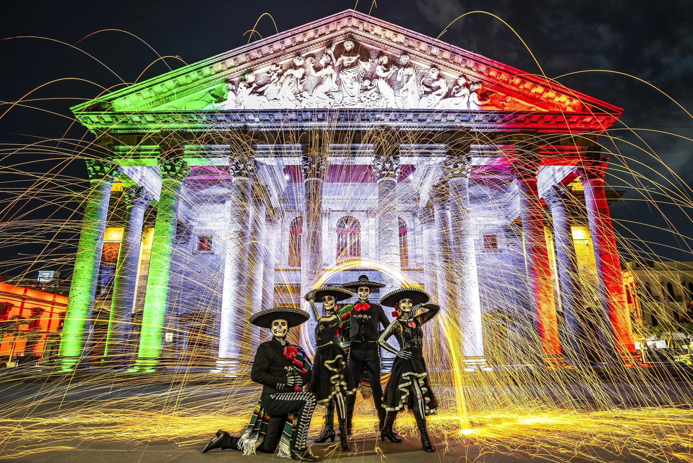 Four people in skeleton costumes pose before a grand building lit in Mexico’s flag colors.