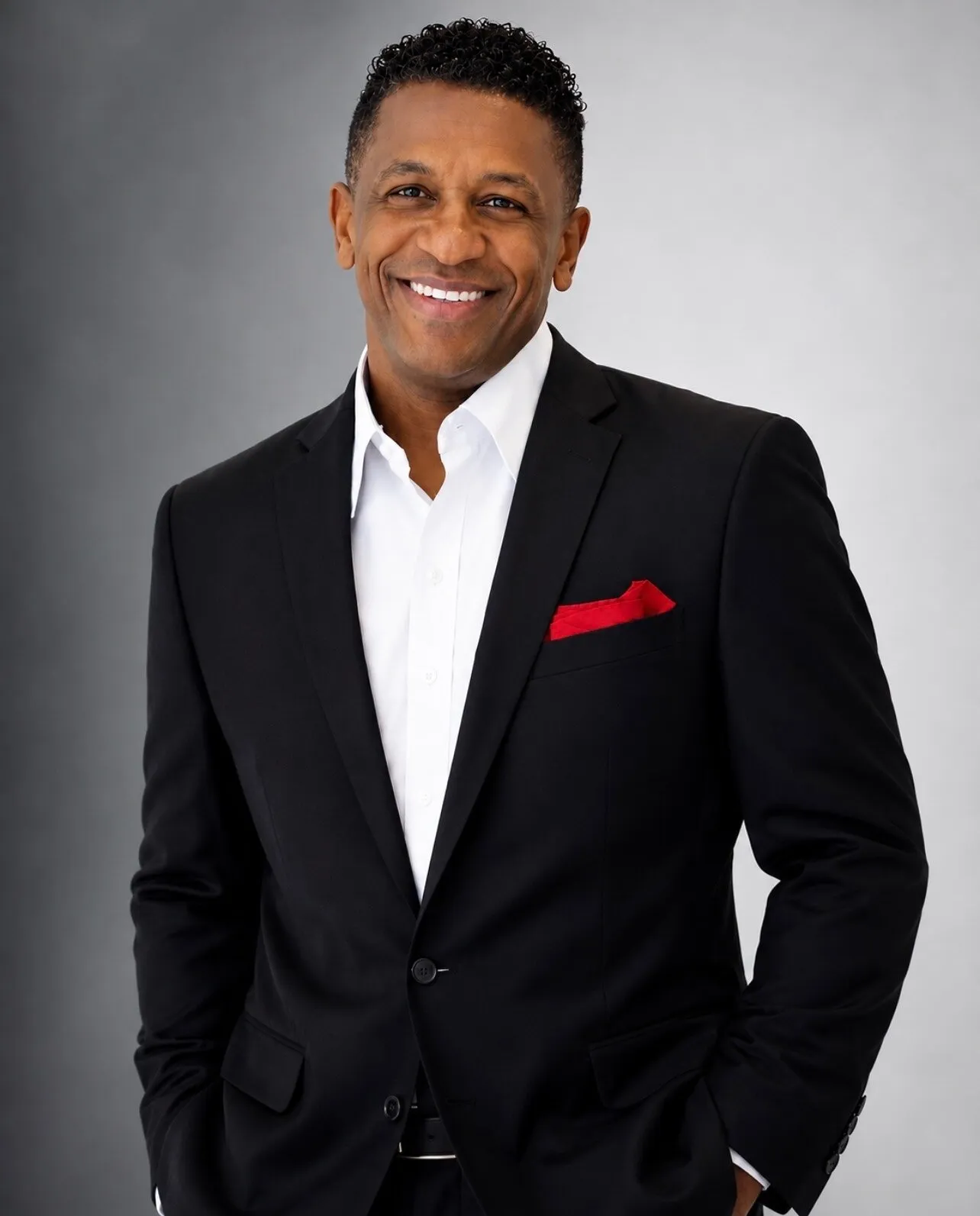 Smiling man in a black suit, white shirt and red pocket square against a gray background.