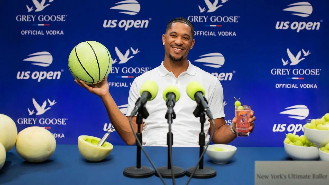 Josh Hart holds a tennis-ball basketball and cocktail behind microphones at a Grey Goose US Open backdrop.