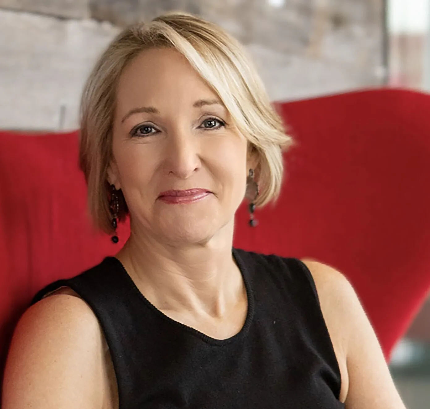 Laurie Pasler wearing a black sleeveless top and dangling earrings against a red background.