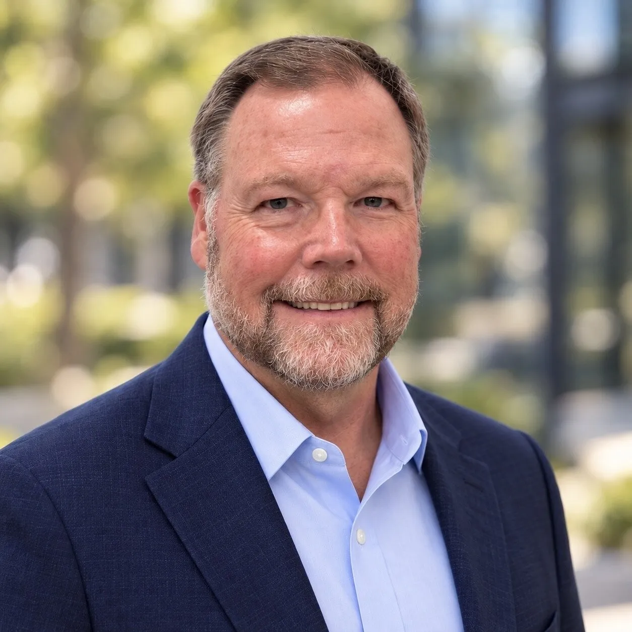Smiling bearded man in a navy blazer and light blue shirt outdoors