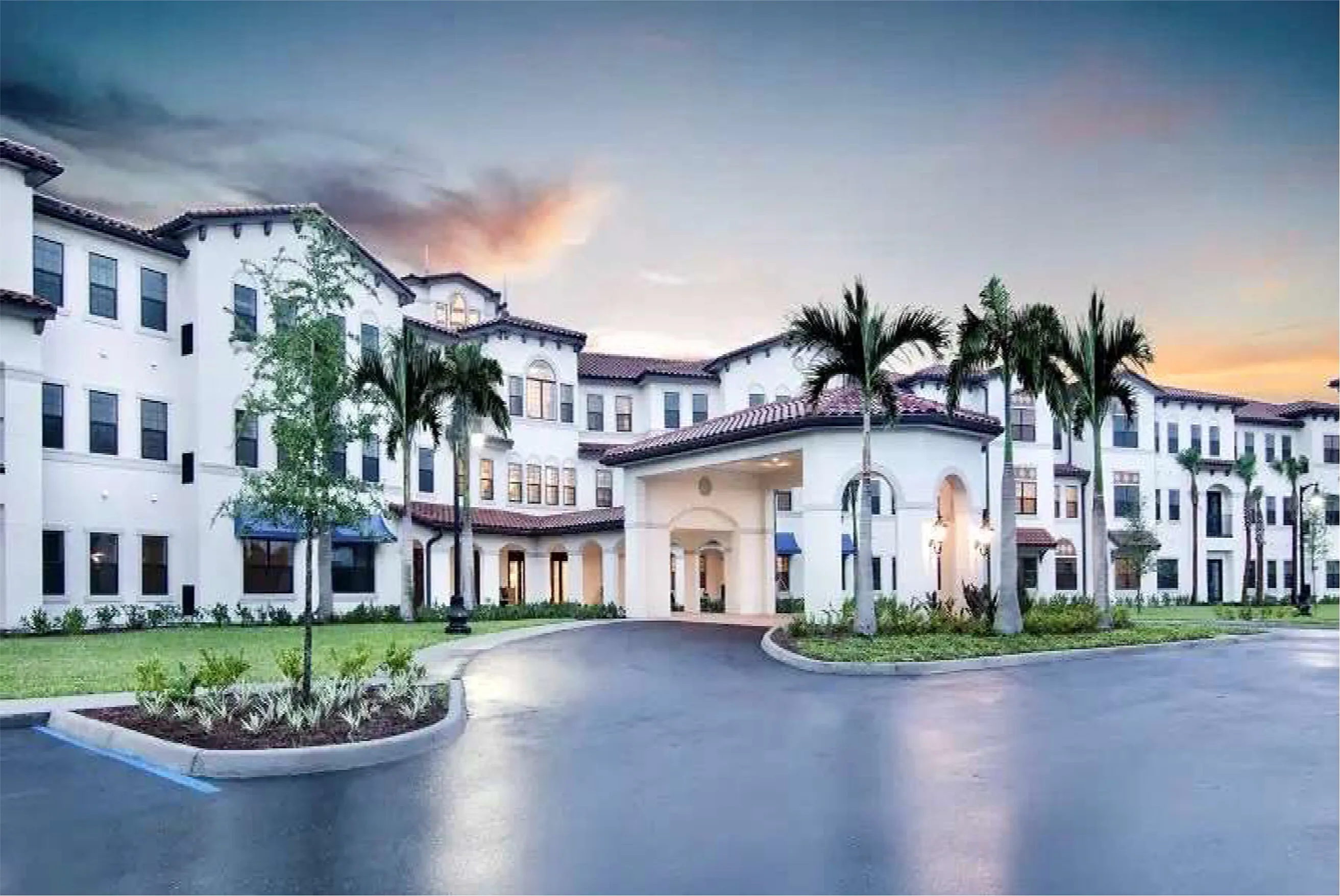White multi-story senior living building with a covered entrance, palm trees and circular driveway at sunset.