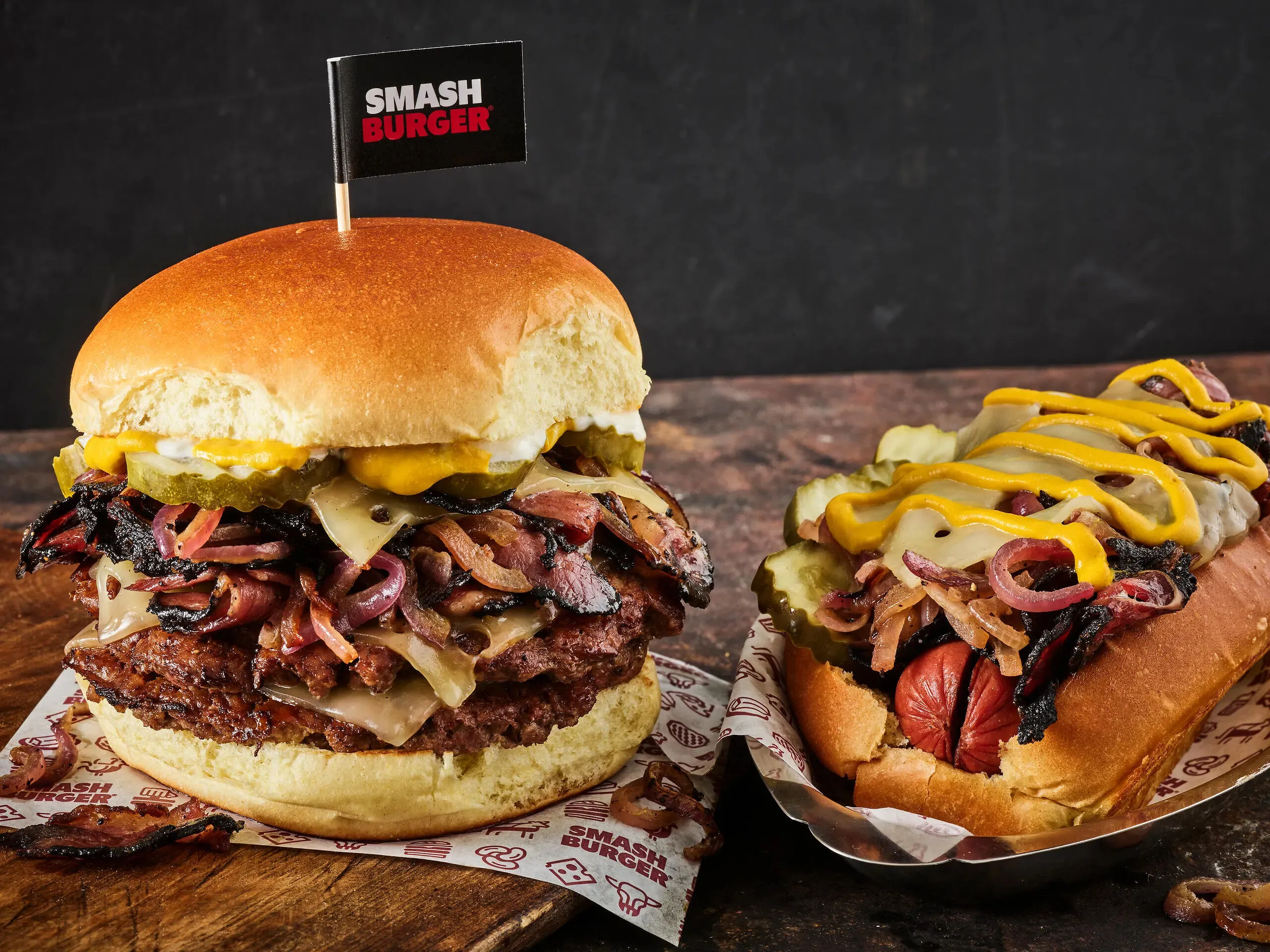 Pastrami-topped cheeseburger beside a hot dog with pickles, onions and mustard.