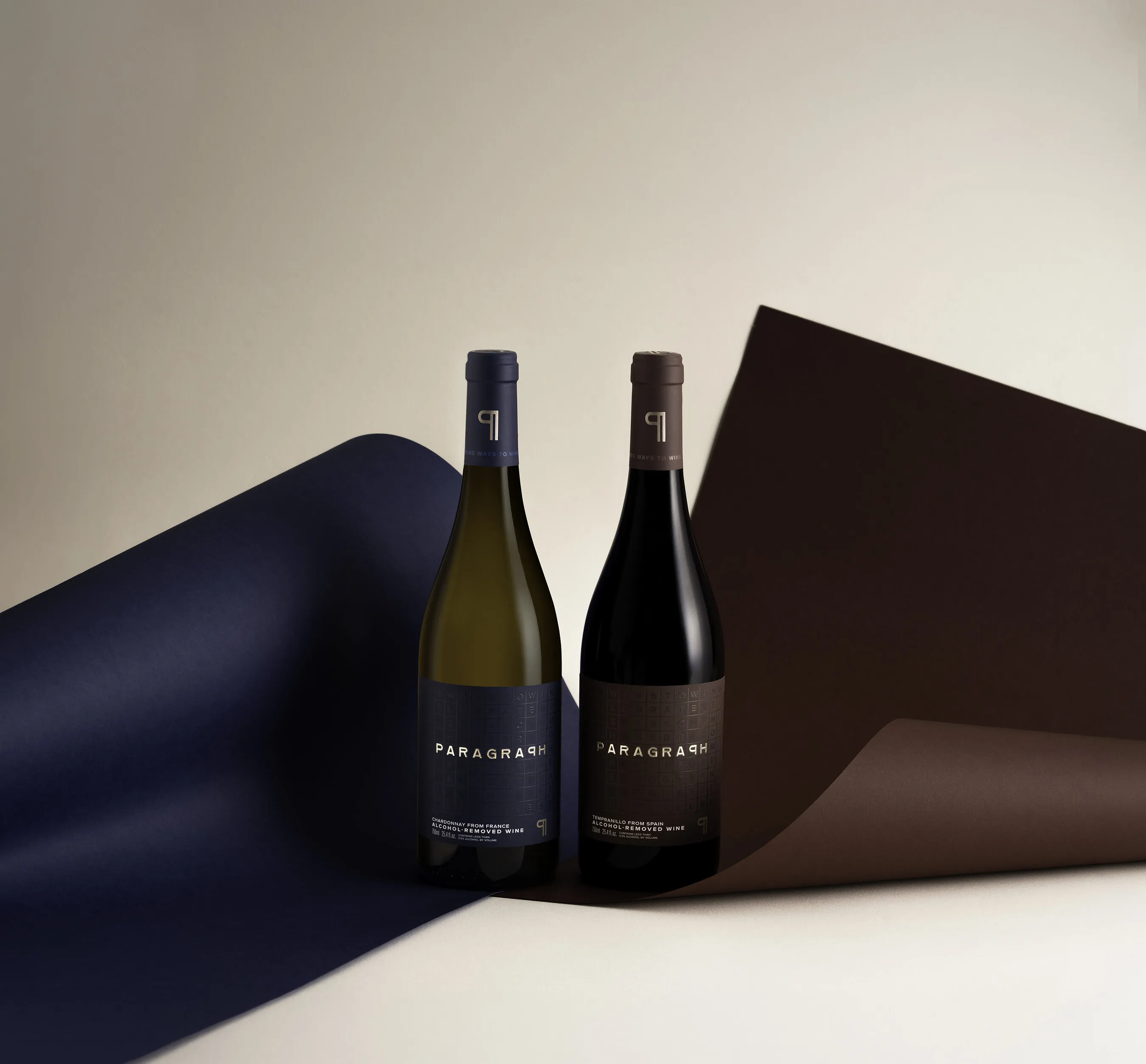 Two PARAGRAPH alcohol-removed wine bottles, Chardonnay and Tempranillo, in front of curled navy and brown paper on a light backdrop.