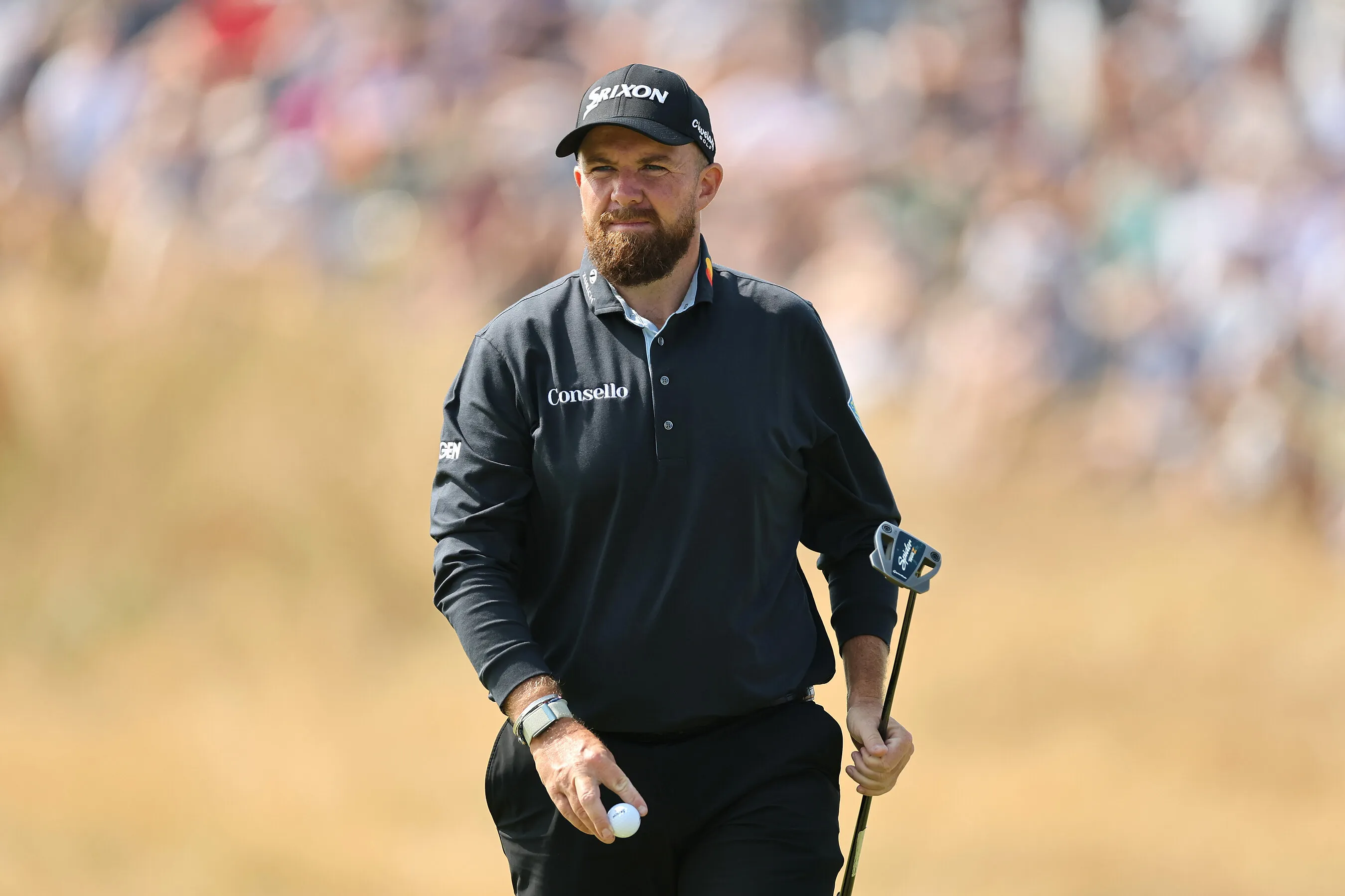 Bearded golfer in a black cap and pullover holding a putter on a course.