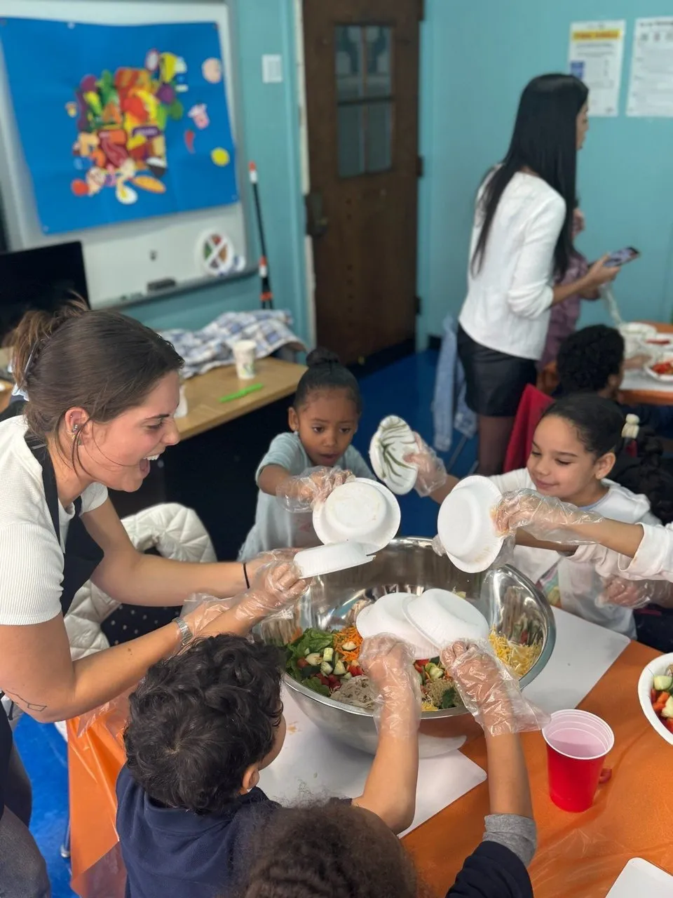 Children and an adult add ingredients to a large bowl during a cooking lesson.