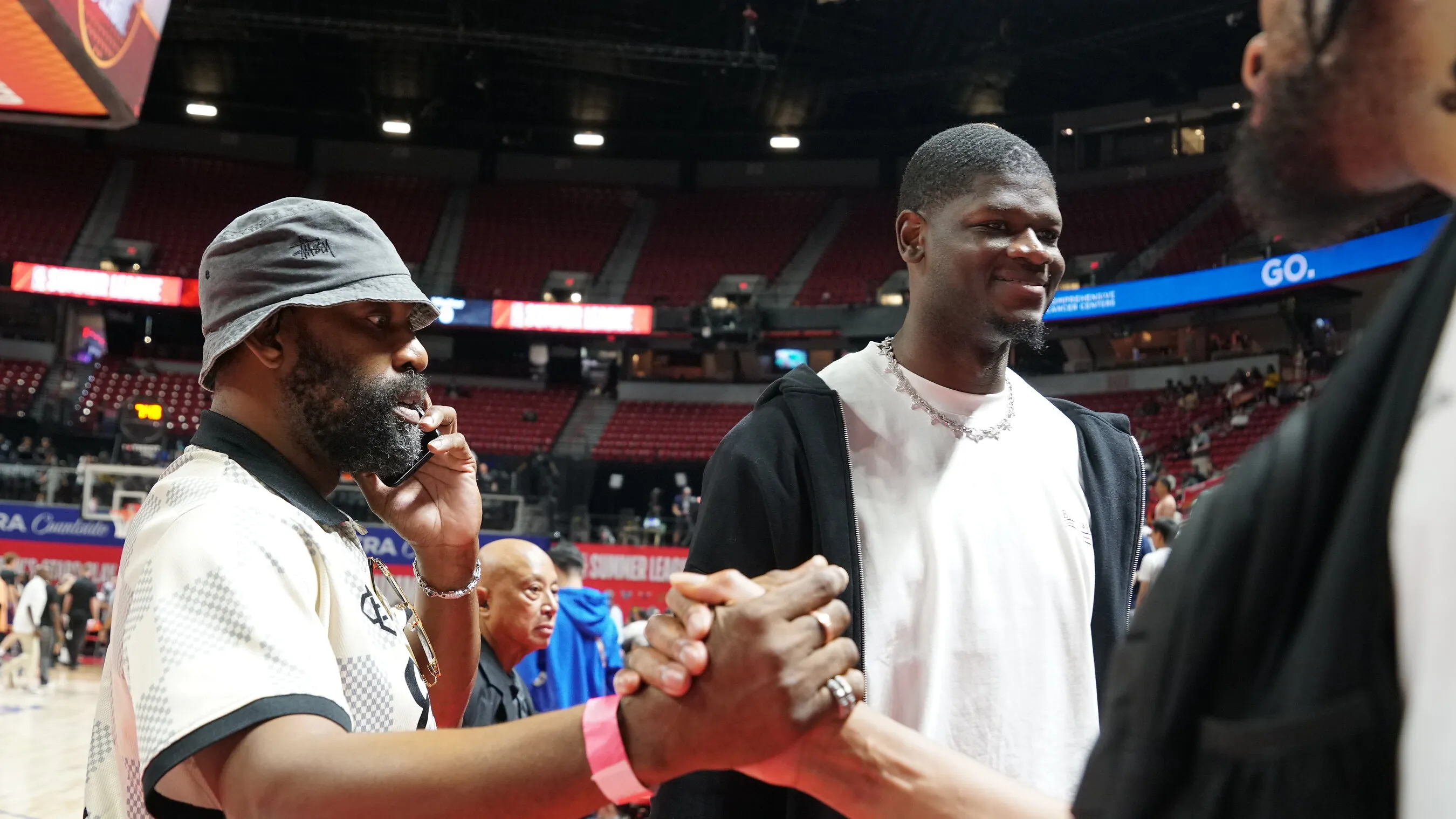 Two men greet each other courtside in a basketball arena while another man speaks on a phone.
