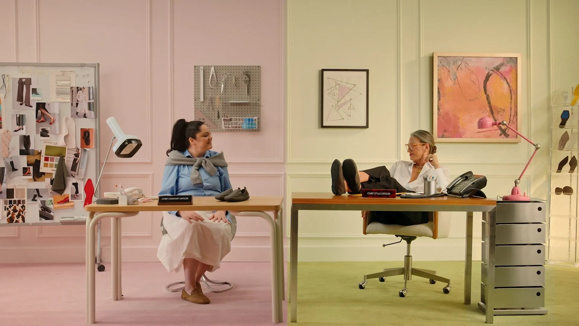 Two women sit at adjoining desks in a pastel shoe design office, with one resting her feet on the desk.