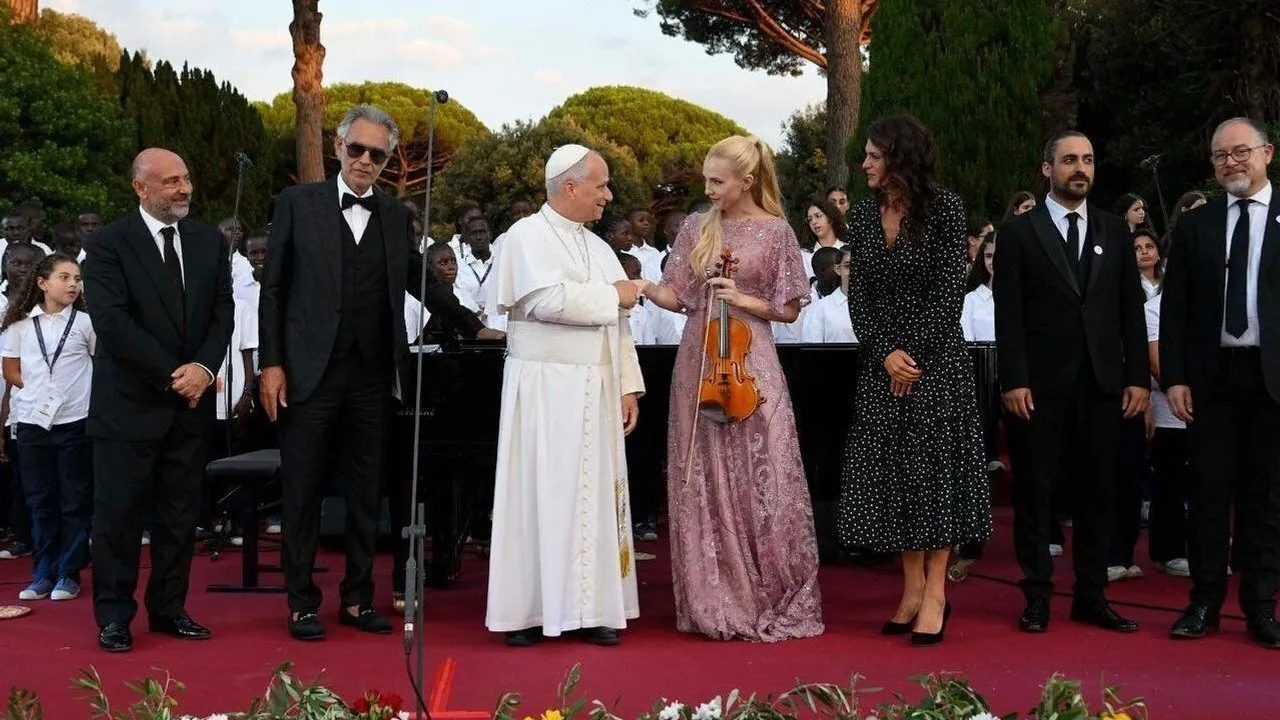 Pope Leo XIV greets violinist in pink gown beside Andrea Bocelli, with choir and guests behind them.