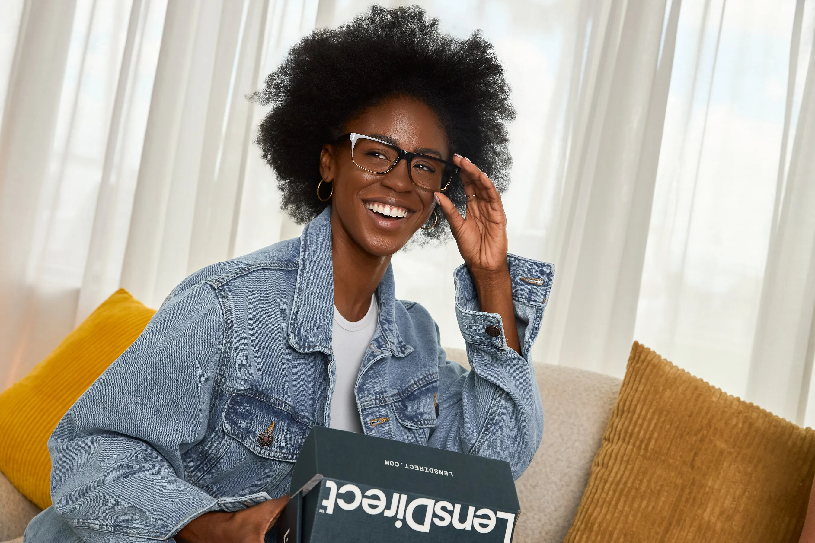 Smiling woman in glasses holding a LensDirect box on a sofa.