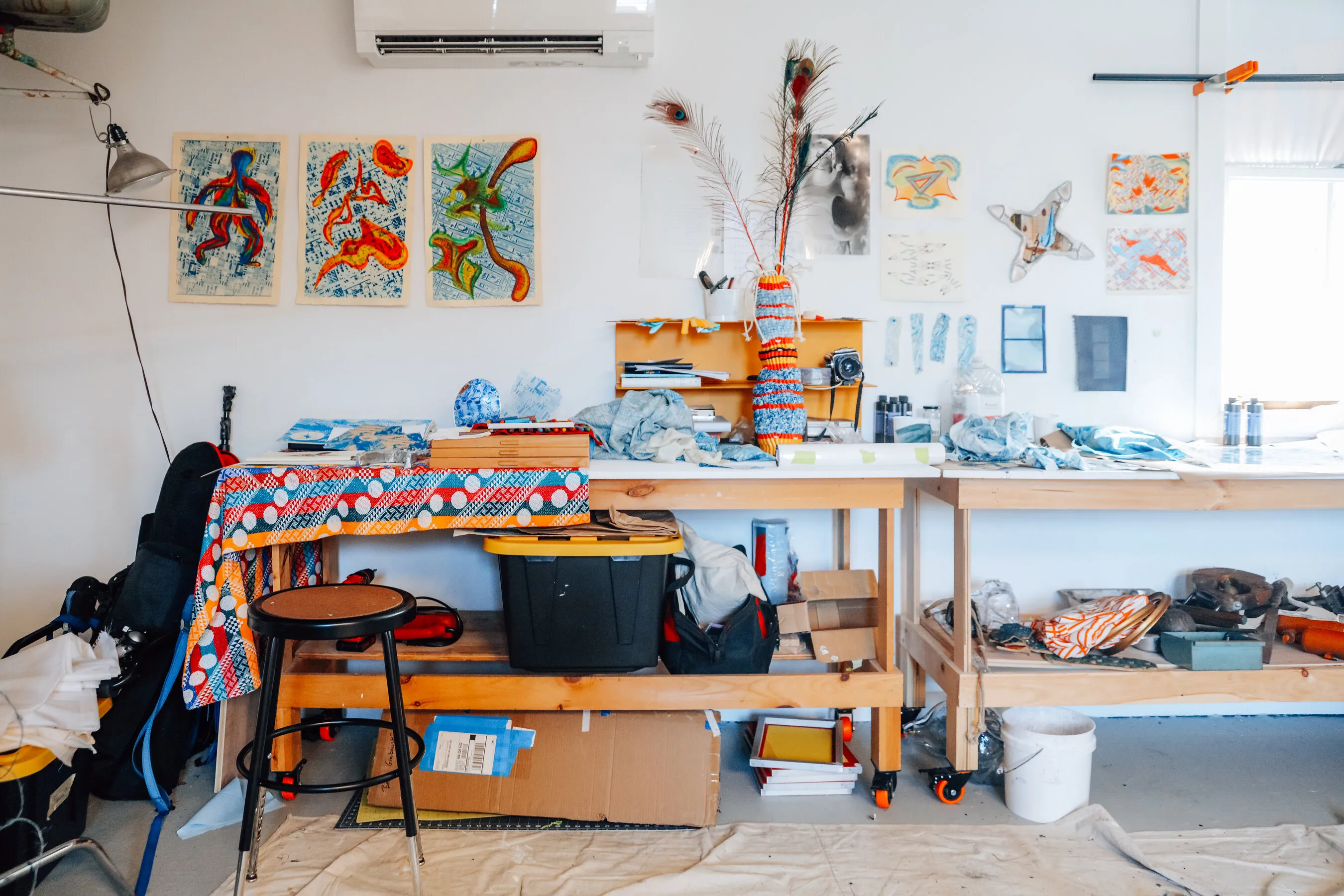 A cluttered art studio with colorful textiles, drawings, worktables and a stool