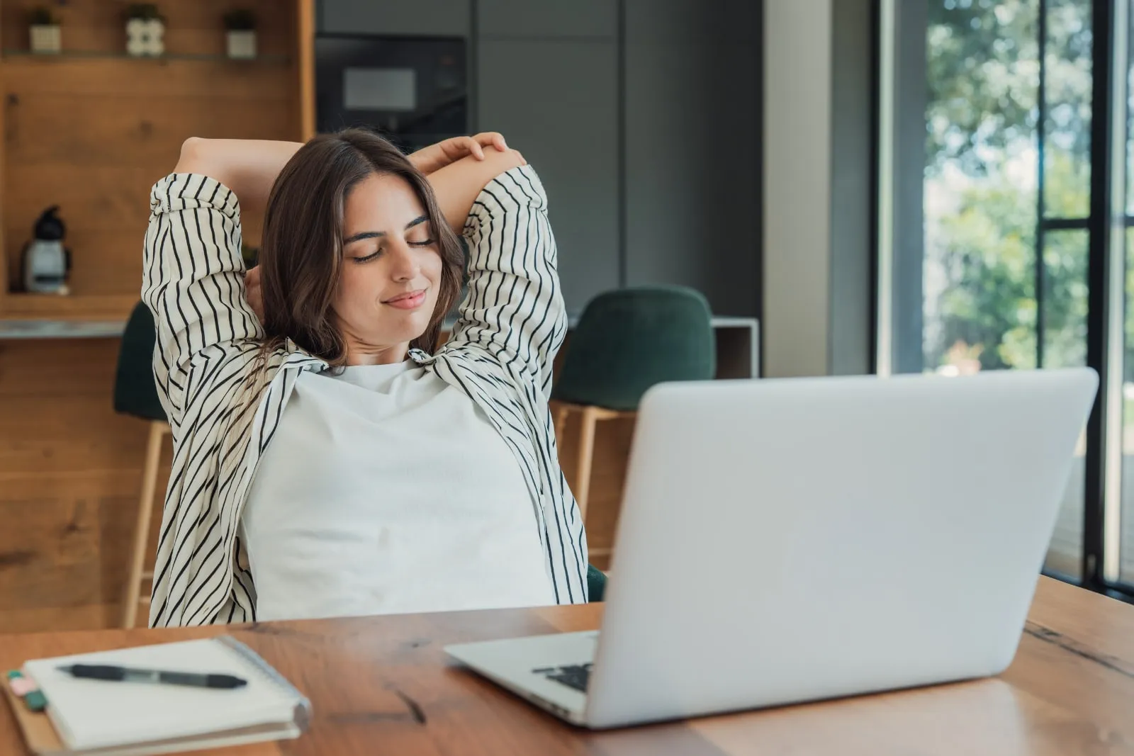 A smiling woman leans back from her laptop with her hands behind her head and her eyes closed.