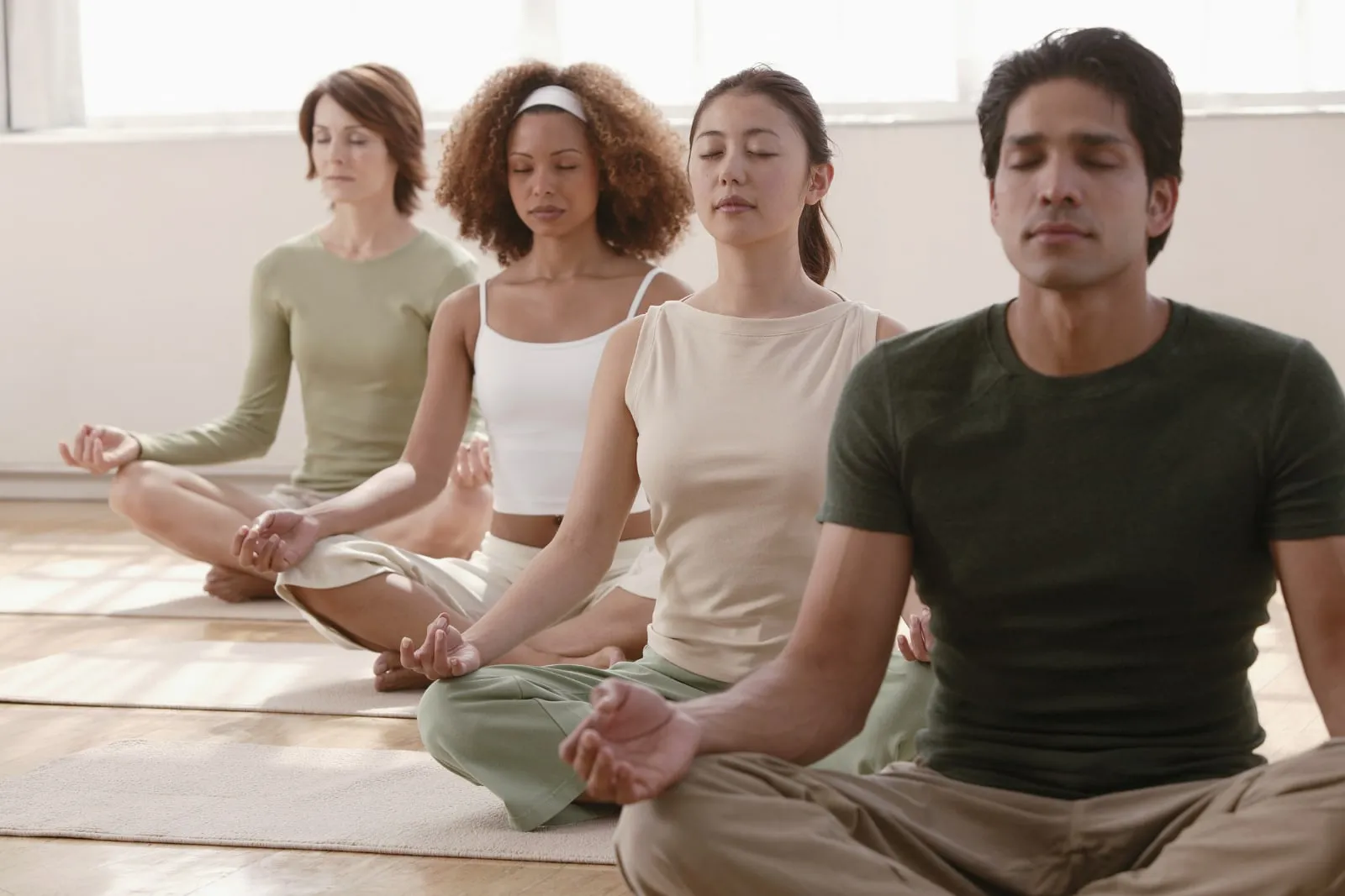 Four people sit cross-legged side by side on a wooden floor in a bright room, eyes closed, hands resting on their knees.