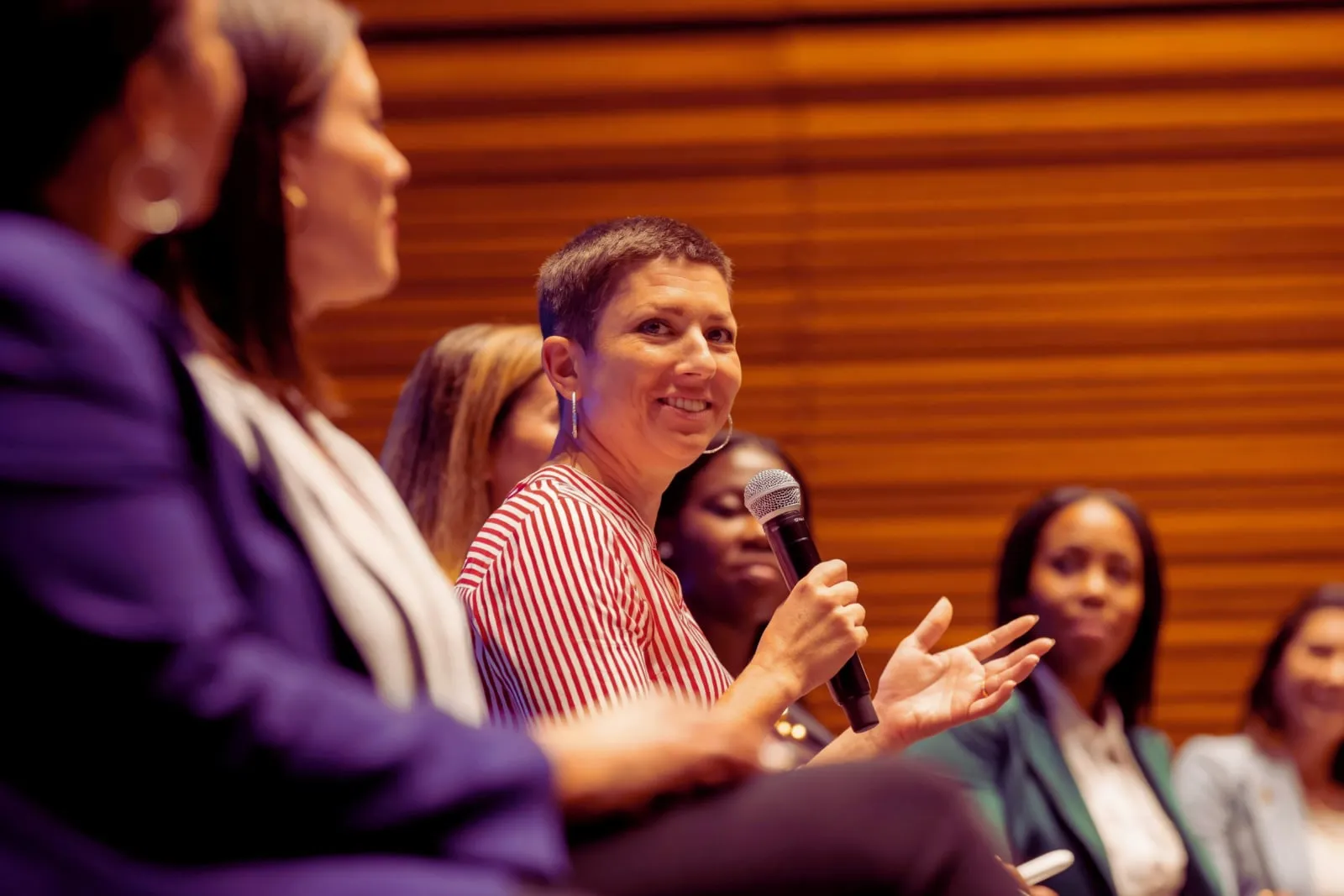 A woman with short hair speaks into a microphone on a panel while other women sit beside her.