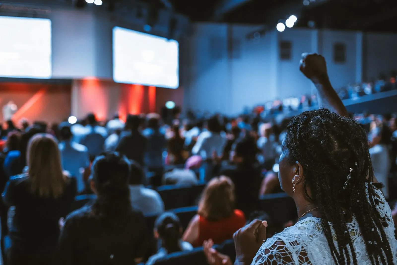 A woman with braided hair sits in a packed auditorium with her fist raised, facing a lit stage.