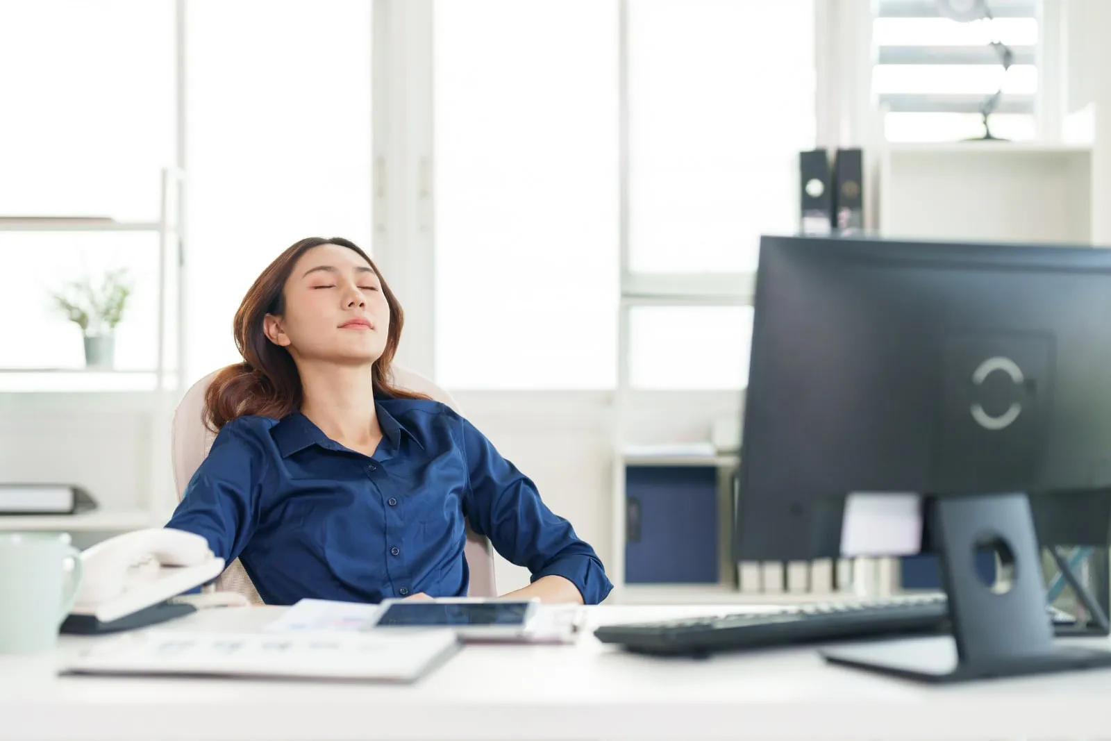 A woman in a blue shirt leans back in her office chair with her eyes closed, a computer monitor and desk beside her.