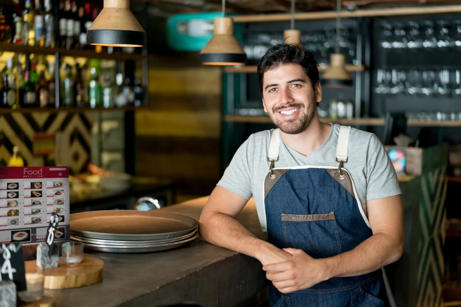 A smiling man in a denim apron leans on the counter of a restaurant.