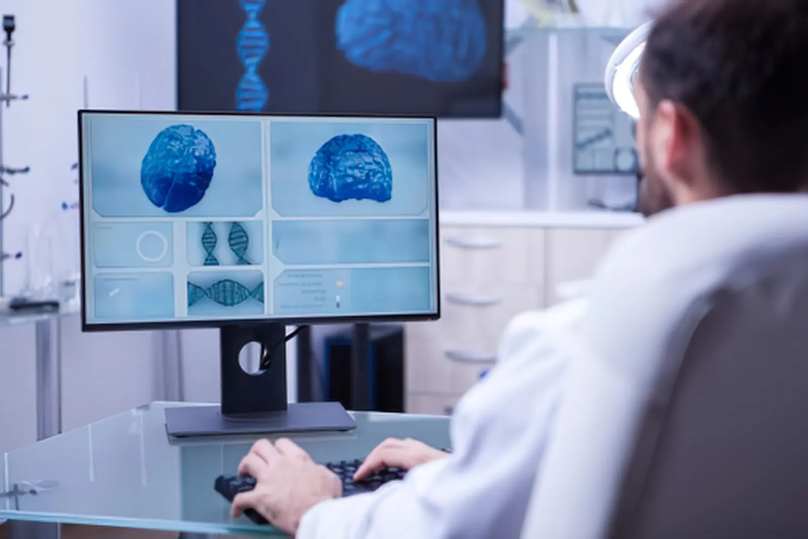 A researcher in a white coat sits at a computer screen showing brain scans and DNA images in a laboratory.