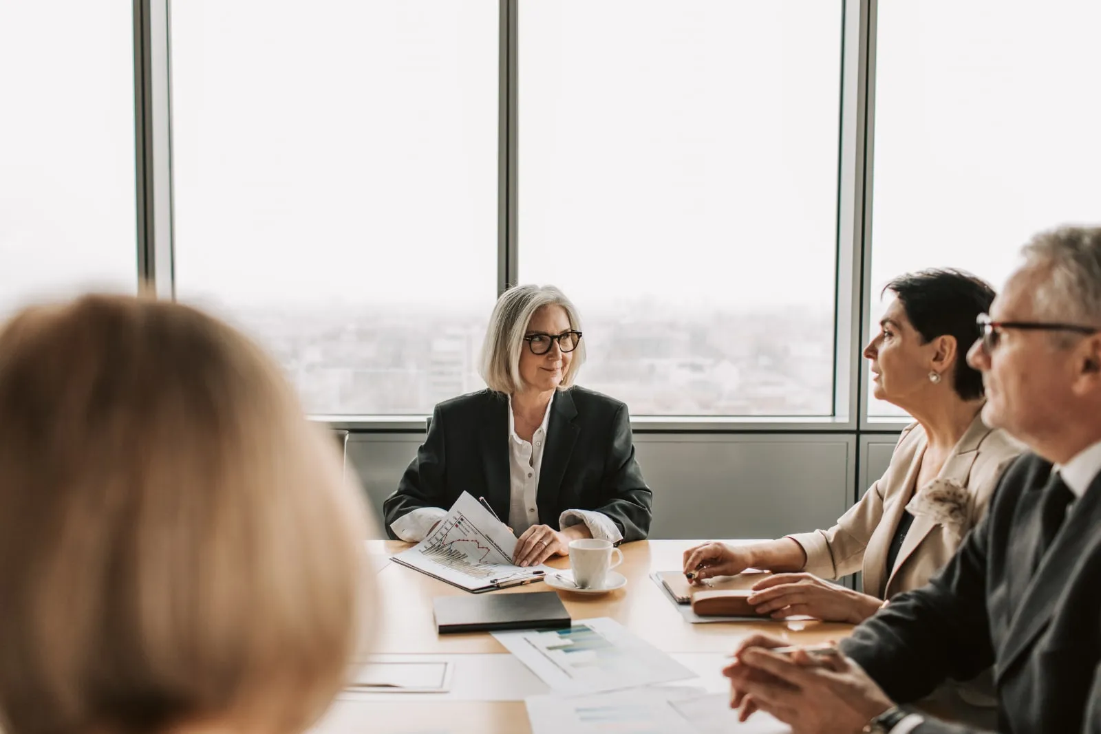 A grey-haired woman in a dark jacket sits at a boardroom table with colleagues, with a view of the city in the windows behind her.