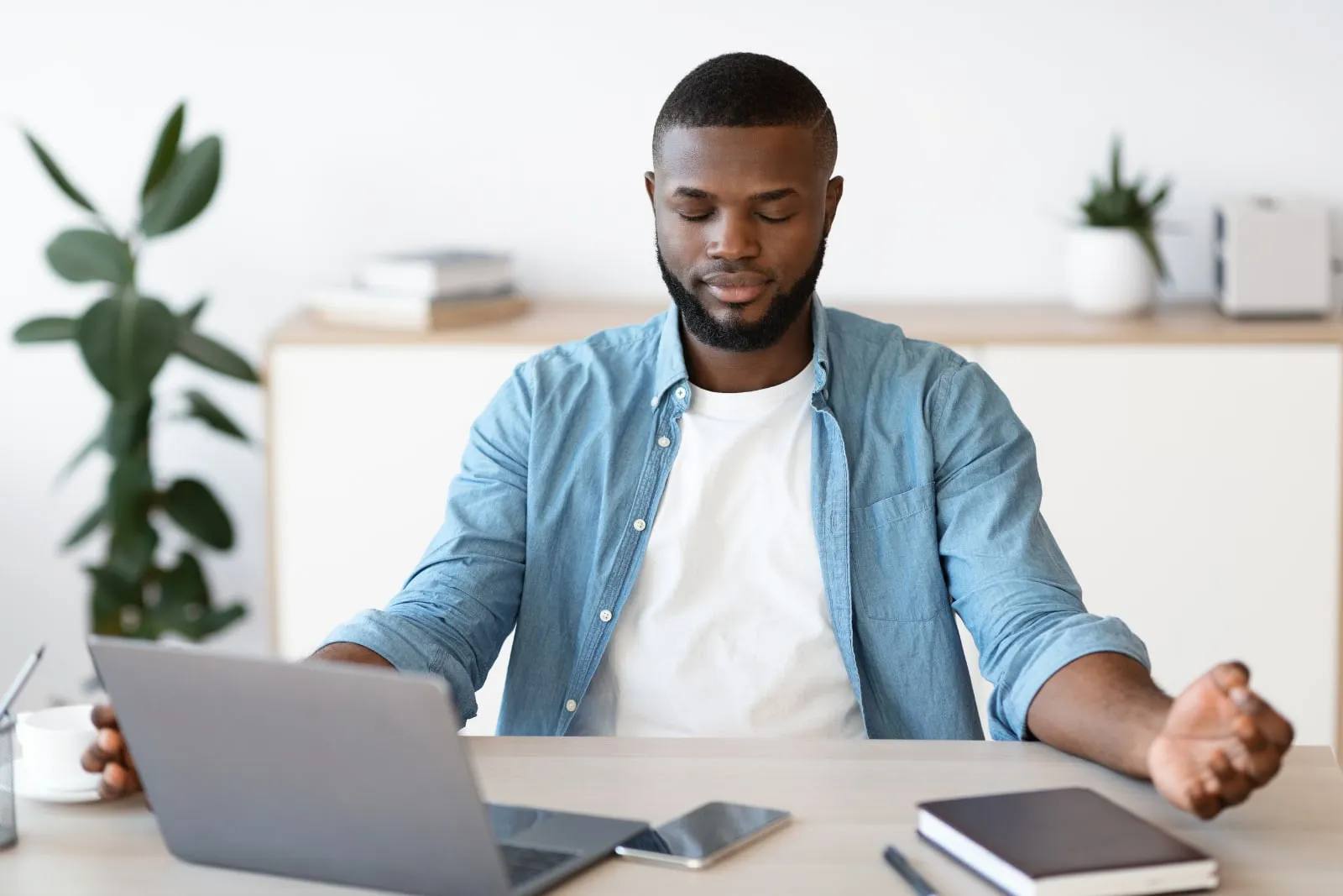A man in a denim shirt sits at a desk with a laptop, his eyes closed and his hands open at his sides.