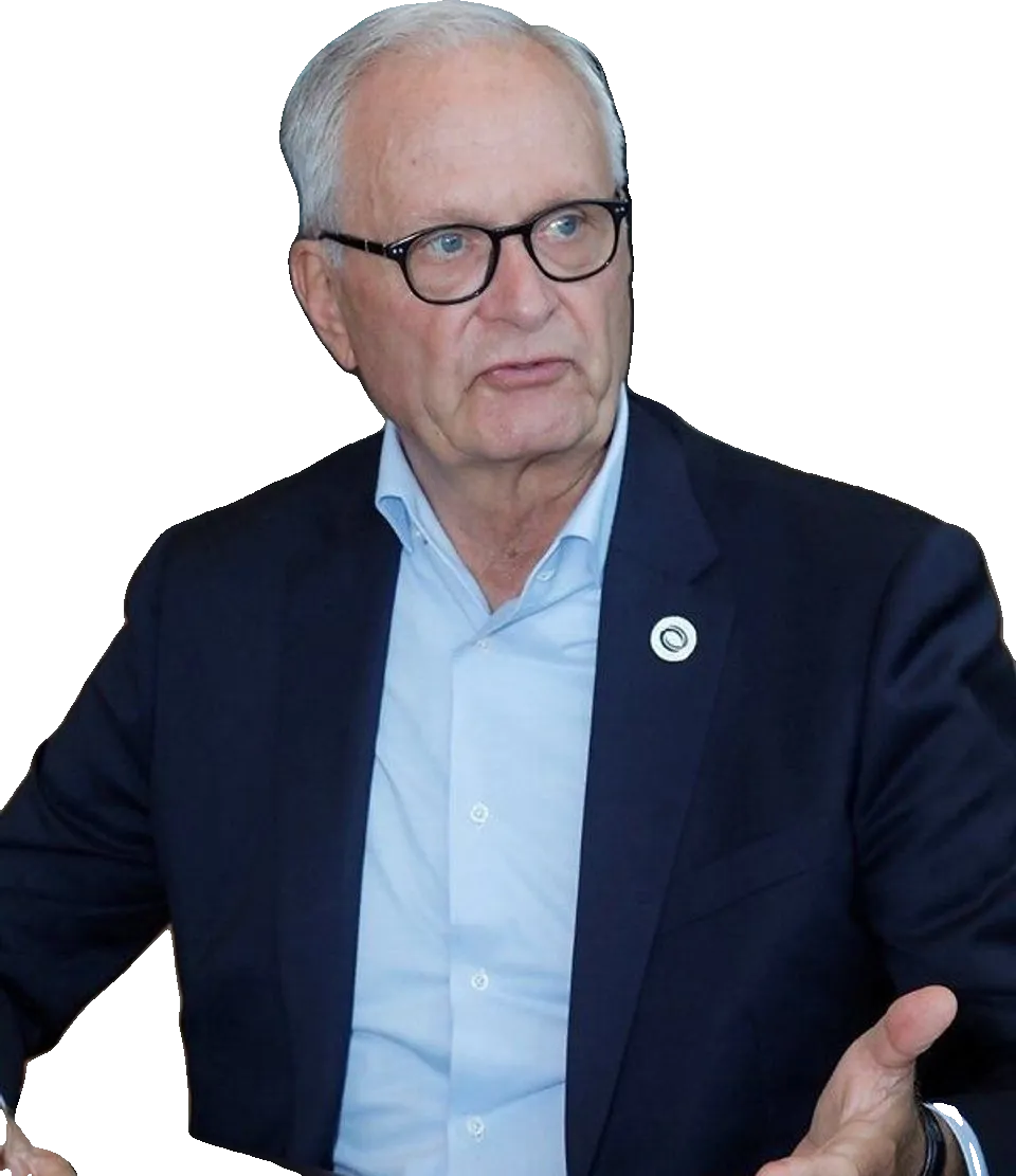 An older man in glasses and a navy suit gestures while seated indoors.
