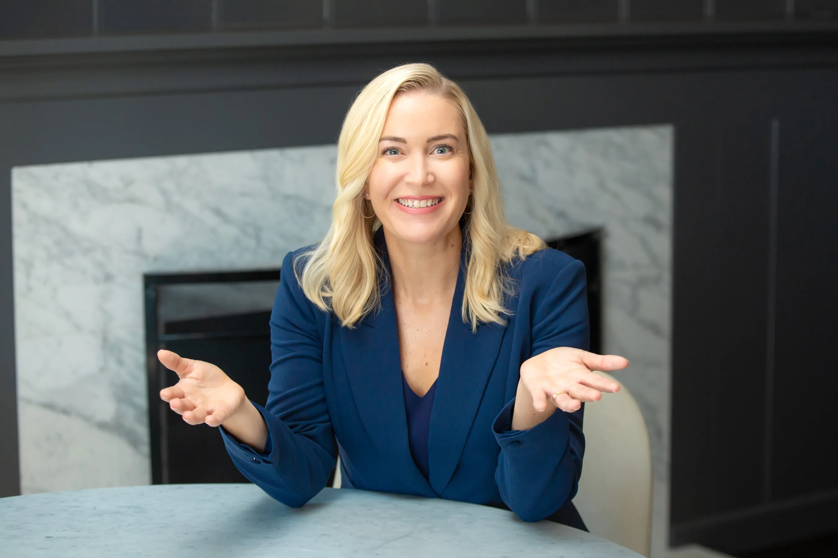 A smiling woman in a blue blazer gestures with open hands in front of a marble fireplace.