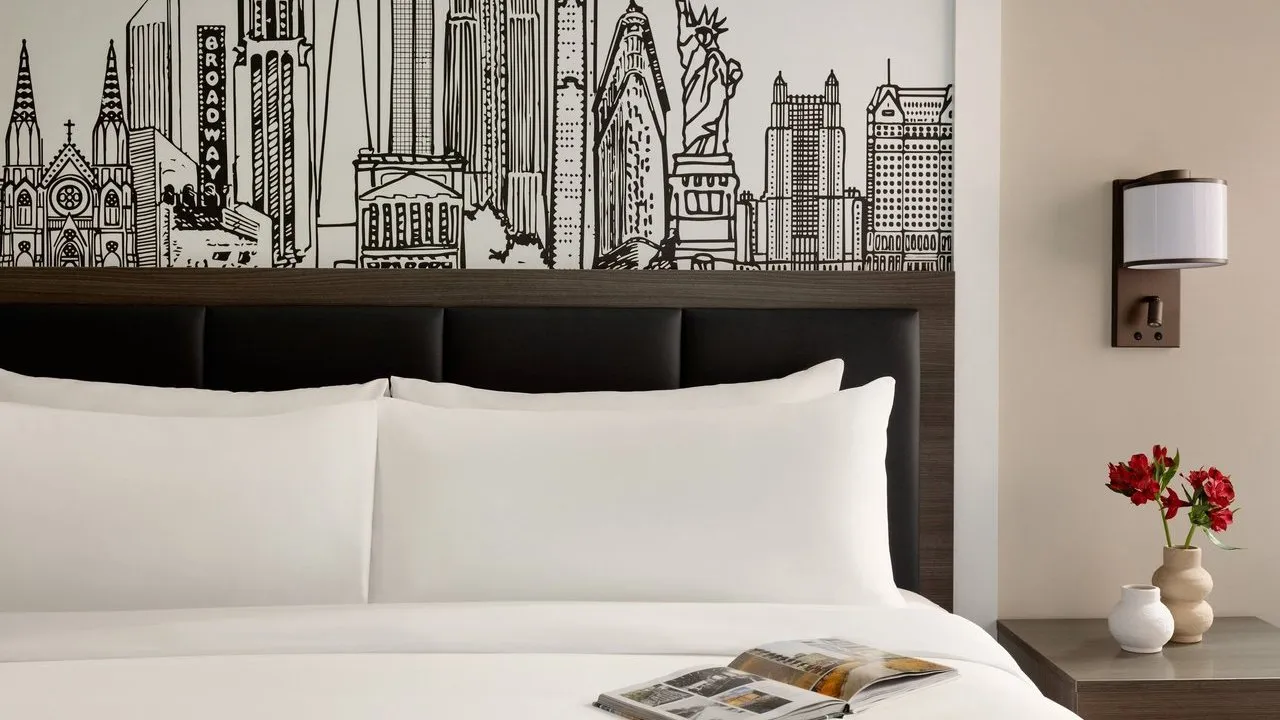King bed with white linens beneath a black-and-white New York skyline mural, beside a lamp and red flowers.