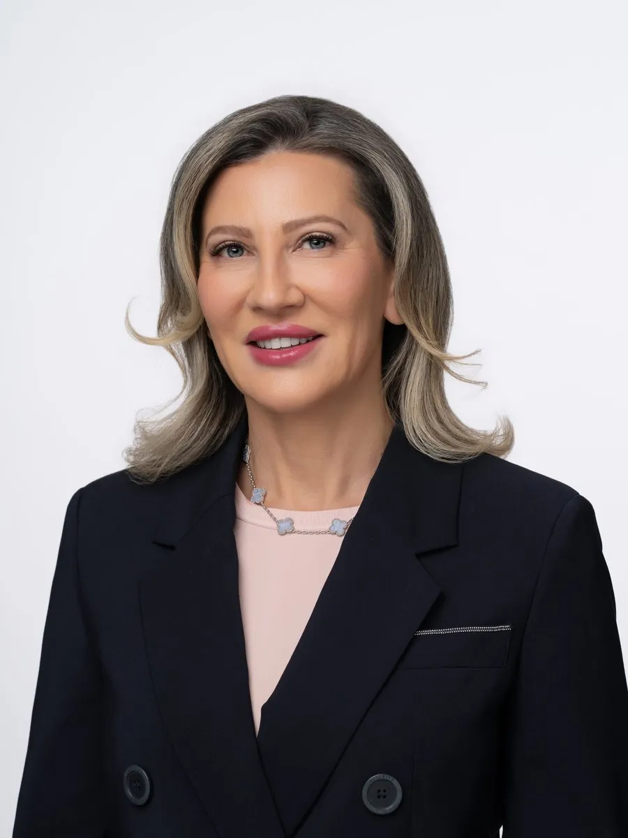 A woman in a dark blazer and pink top against a light background