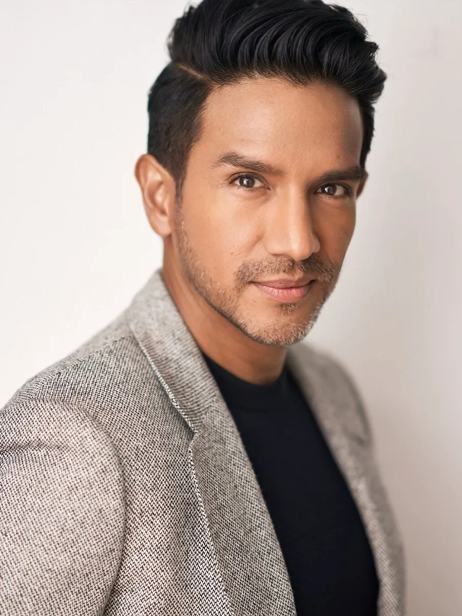 A man with styled dark hair and stubble wearing a gray blazer over a black shirt.