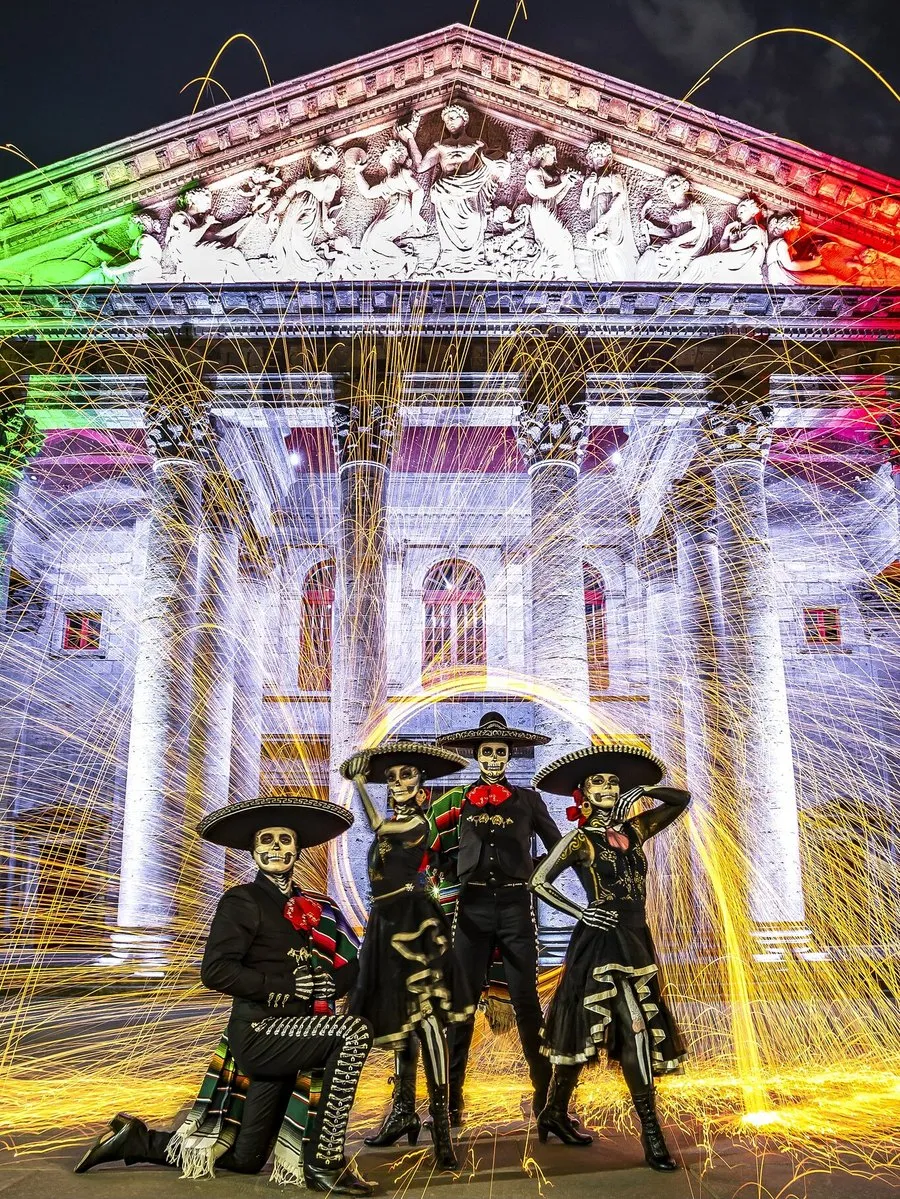Four people in skeleton costumes pose before a grand building lit in Mexico’s flag colors.
